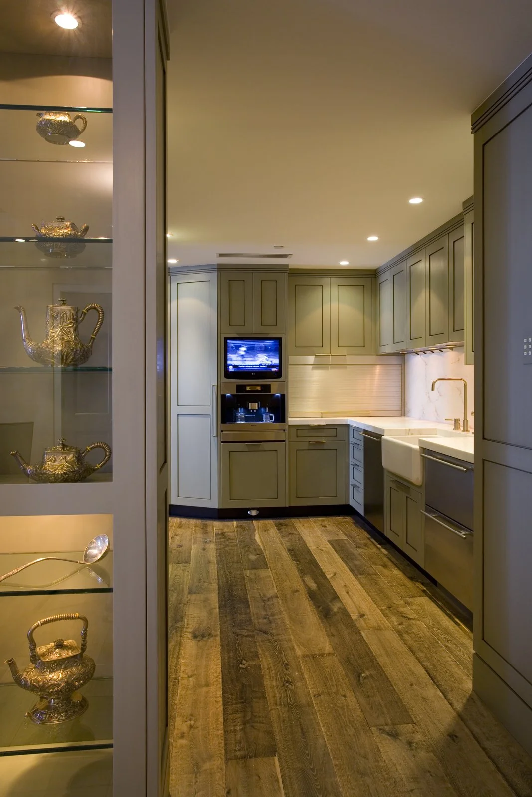 A kitchen with green cabinetry, a white farmhouse sink, a small television, and wooden flooring. There's a glass cabinet on the left displaying gold teapots.