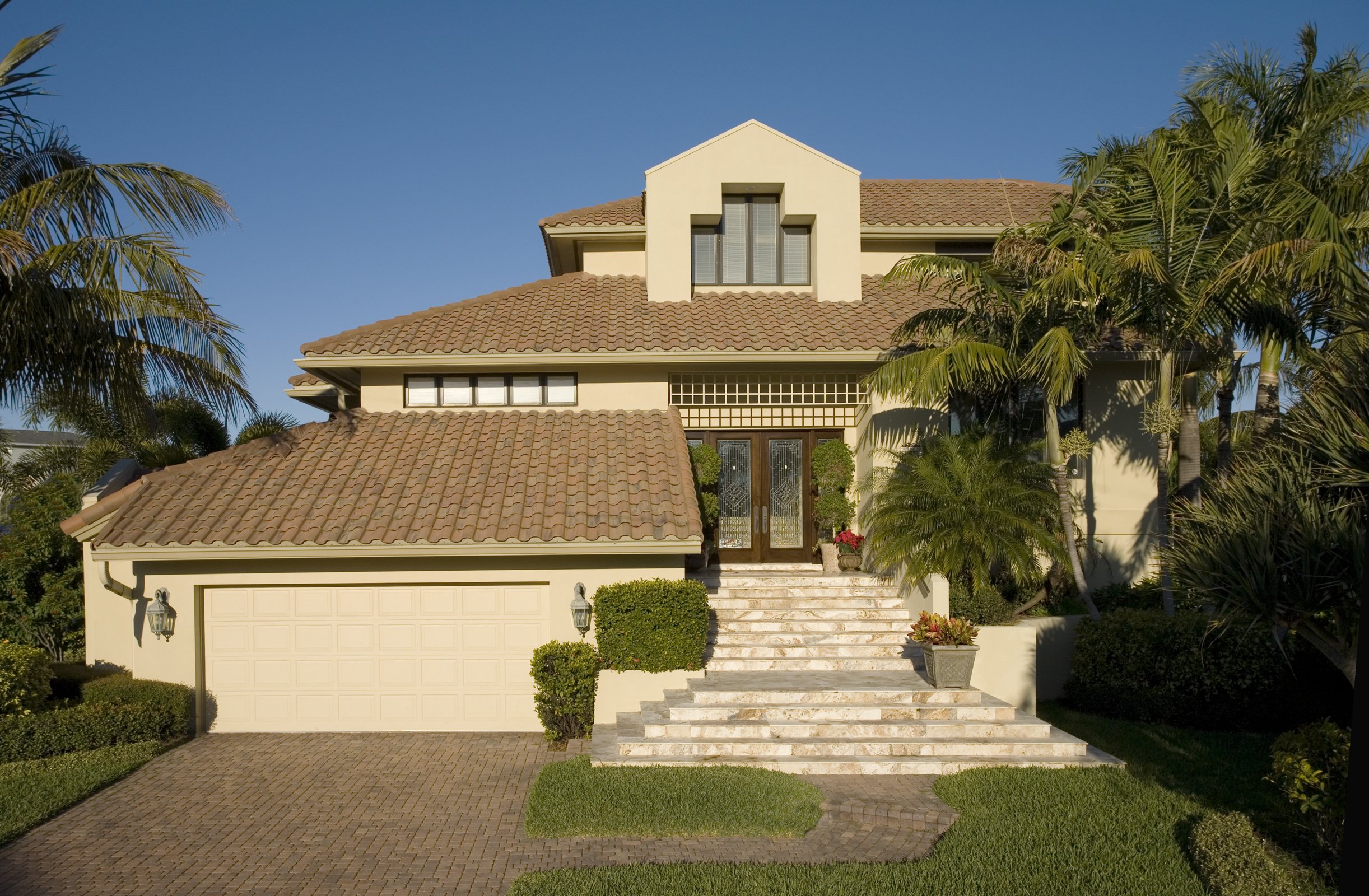 Front view of a large, two-story house with beige walls, brown tiled roof, and a garage. The house features stairs leading to the entrance with decorative plants and palm trees around.