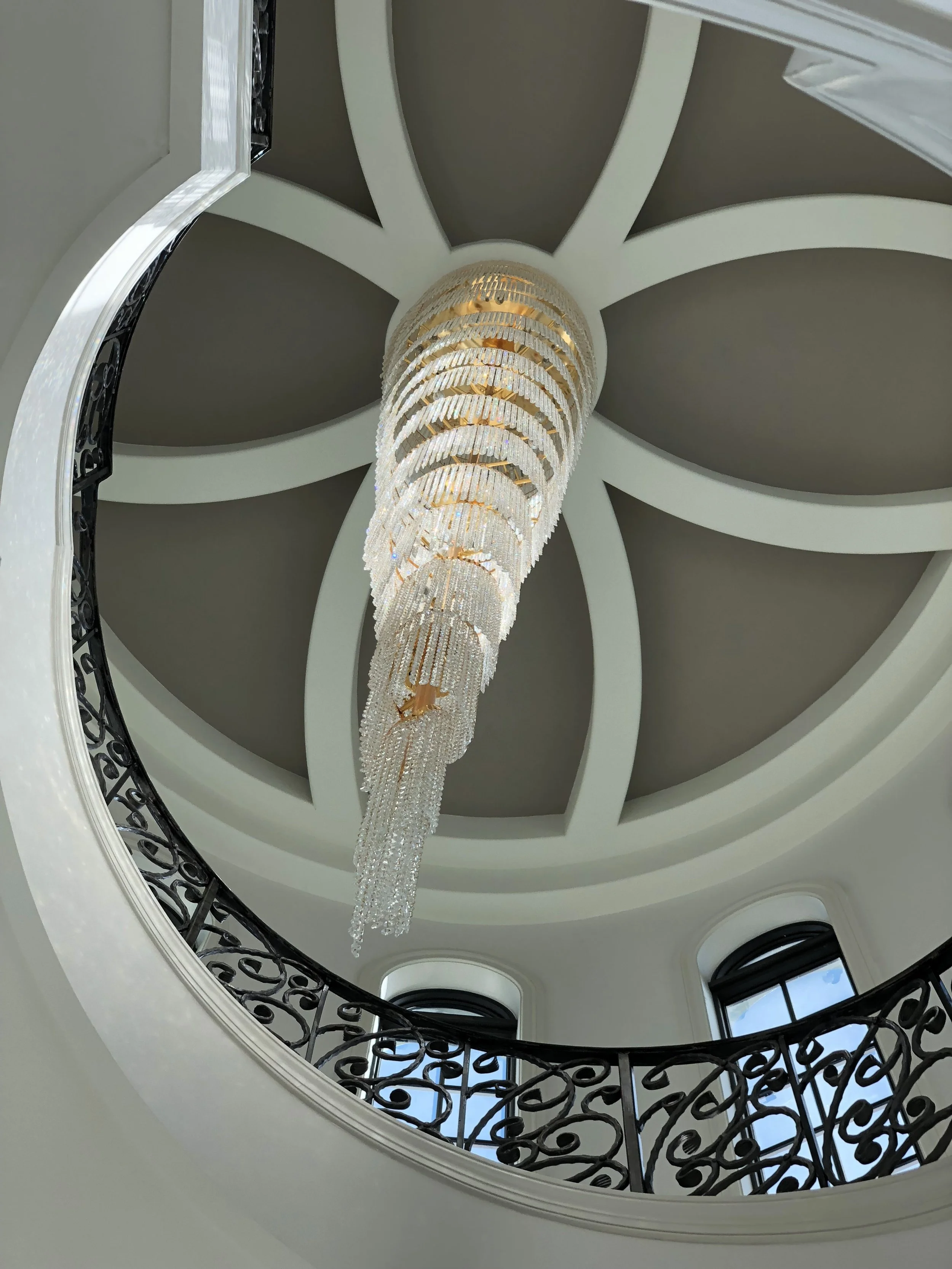Looking up at a grand crystal chandelier hanging from the ceiling of a spiral staircase with decorative black wrought iron railing and large arched windows.