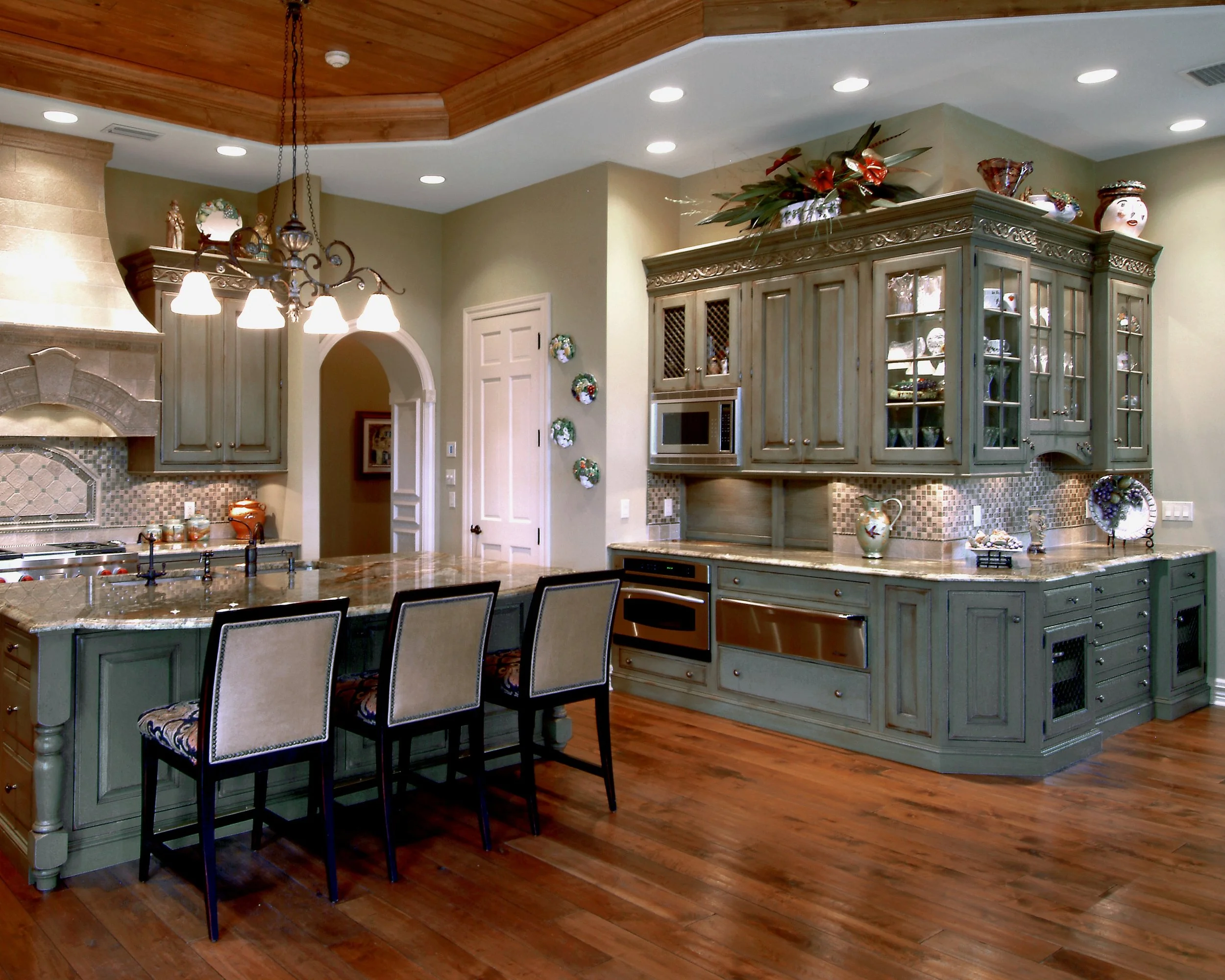 A spacious kitchen with green cabinetry, a marble island, wood flooring, and a decorative ceiling with a chandelier.