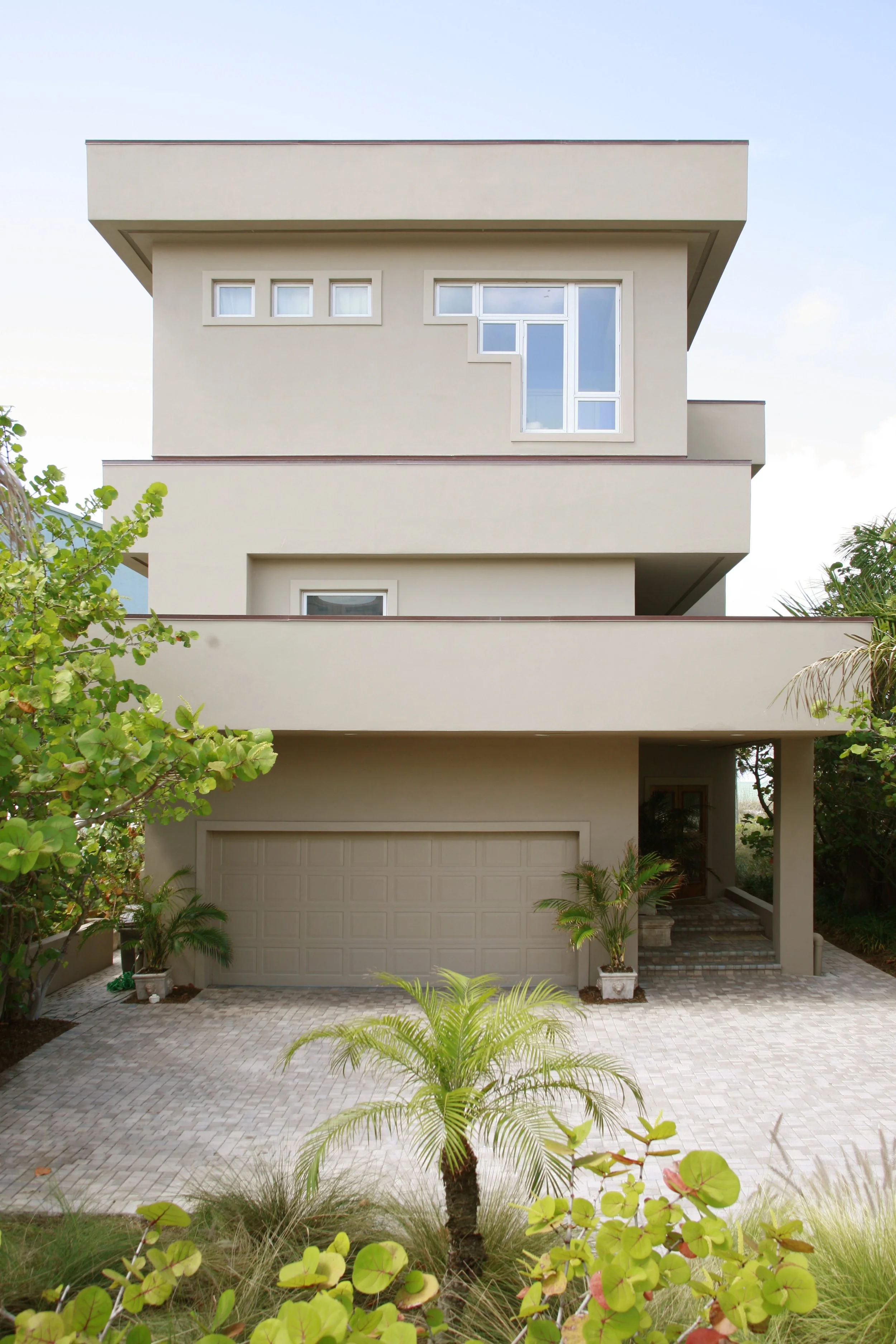 A modern three-story house with balconies, beige exterior, large windows, a garage, and tropical plants in front.