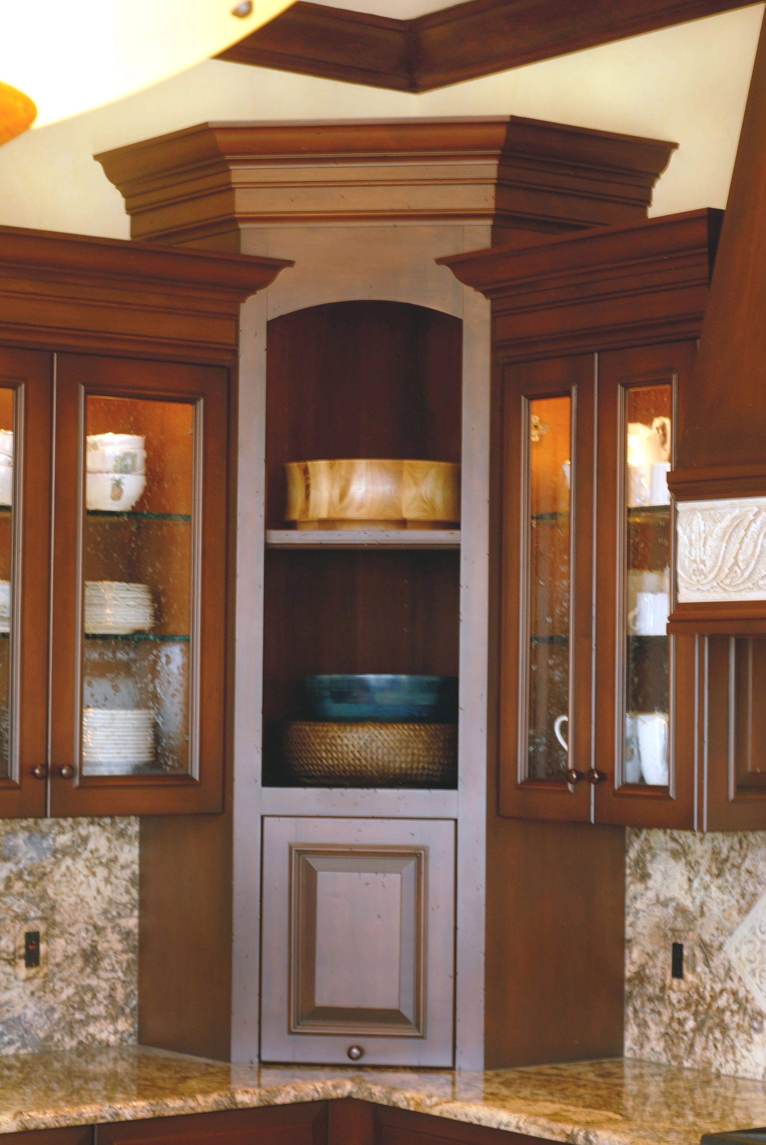 A wooden kitchen cabinet with glass doors, displaying dishes, and an open central section with shelves containing bowls, with a granite countertop below and a decorative wooden top above.
