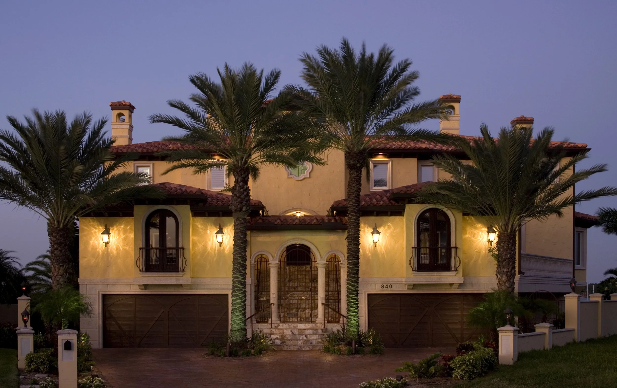 A large Mediterranean-style house illuminated at dusk with palm trees in front, yellow stucco walls, arched windows, wrought iron balcony, and a gated entrance.