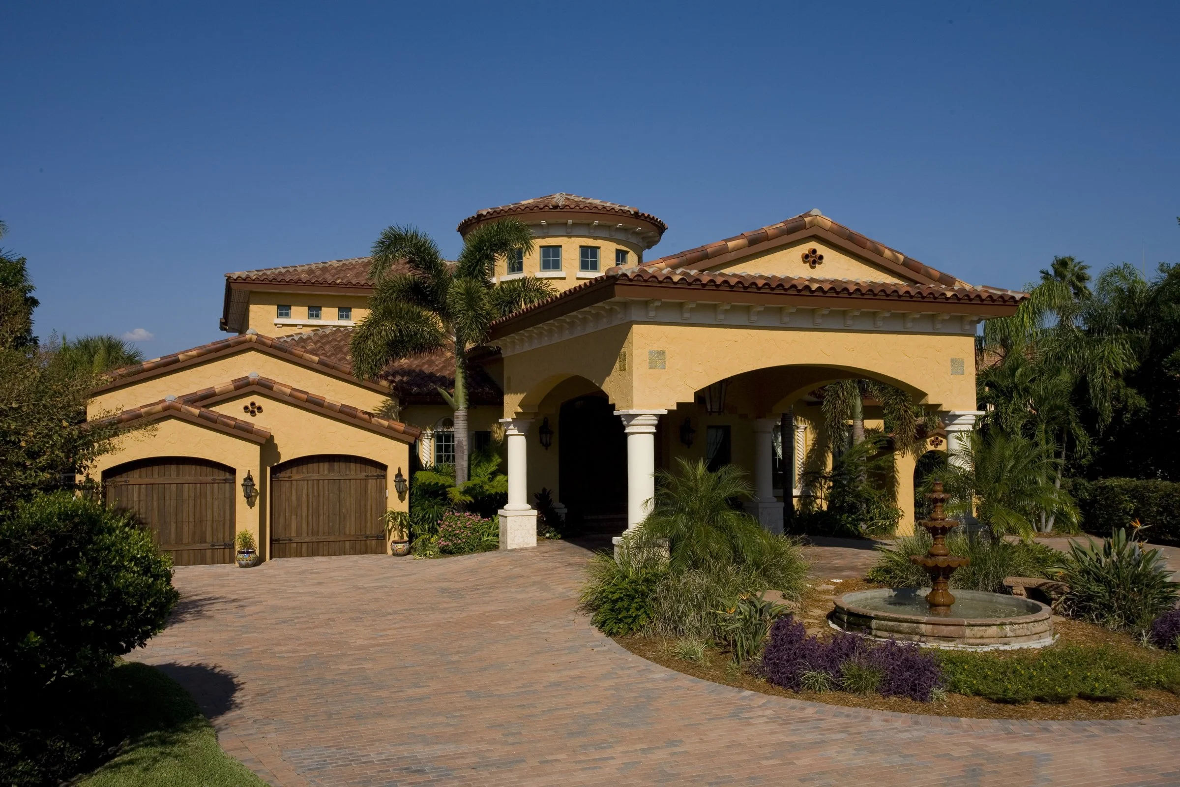A large, yellow, Mediterranean-style house with red-tile roof, surrounded by lush landscaping, including palm trees and a water fountain in the front yard.