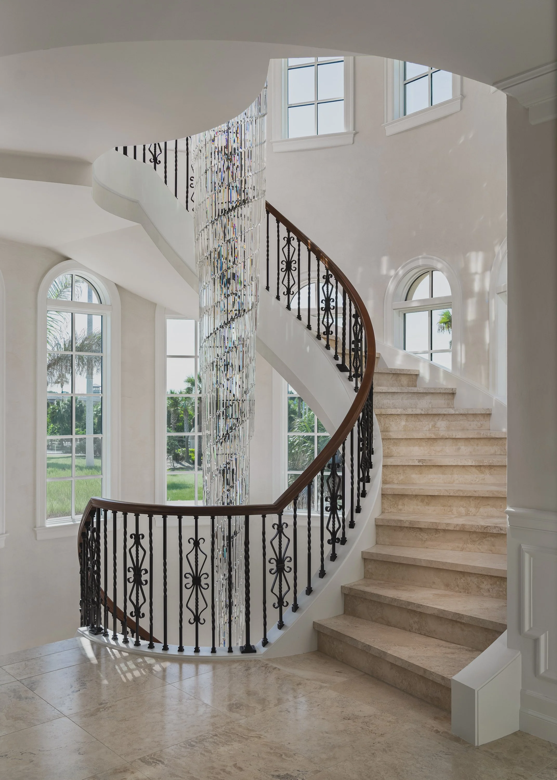 Interior view of a grand staircase with beige marble steps, black wrought iron railing with decorative patterns, wooden handrail, and a shimmering crystal chandelier hanging from the ceiling. Large arched windows with white trim allow natural light to fill the space, revealing a green outdoor landscape.