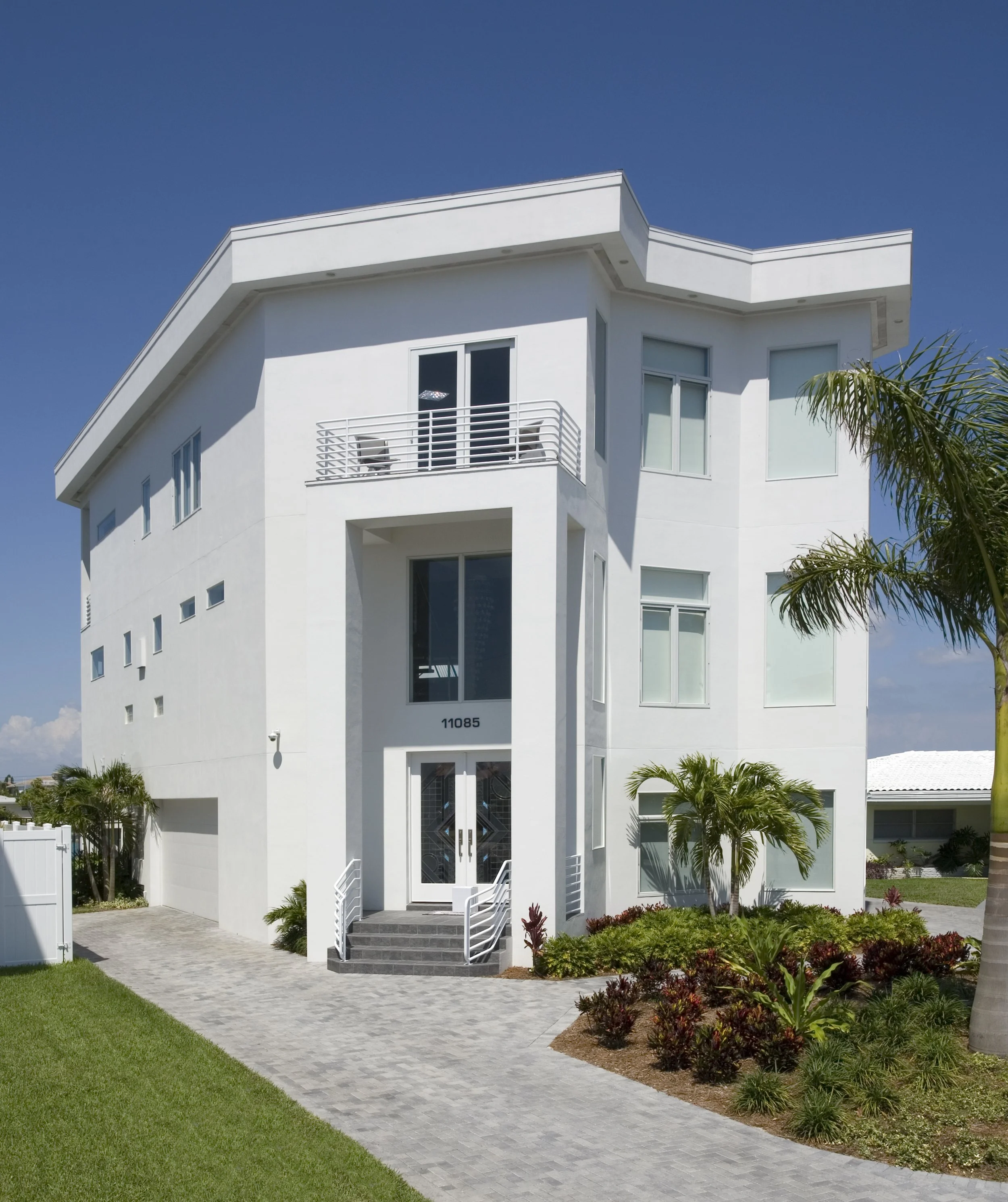 Modern white multi-story residential building with a landscaped front yard, palm trees, and a paved walkway under a clear blue sky.