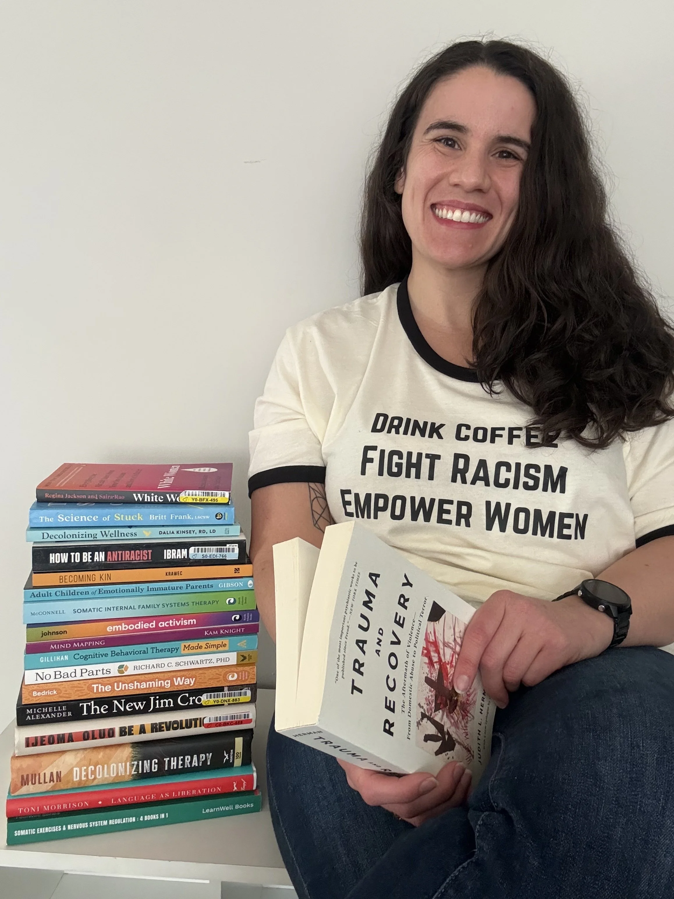 Amanda Kenney, LCSW sitting holding Trauma and Recovery by Judith Herman. Next to her is a pile of various books, with themes of therapy and social justice.