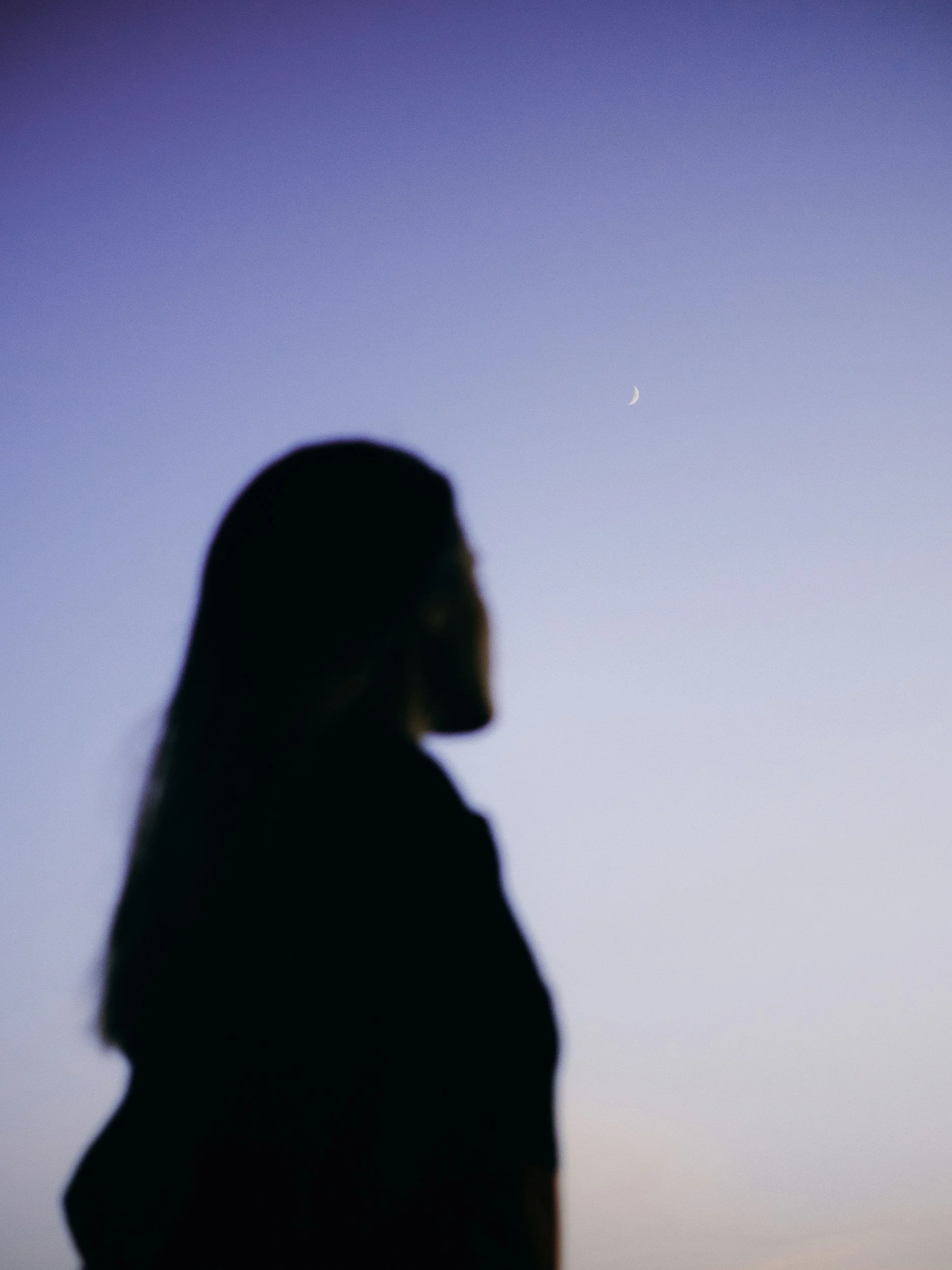 Silhouette of a woman with long hair looking towards the sky with a crescent moon and a faint star.