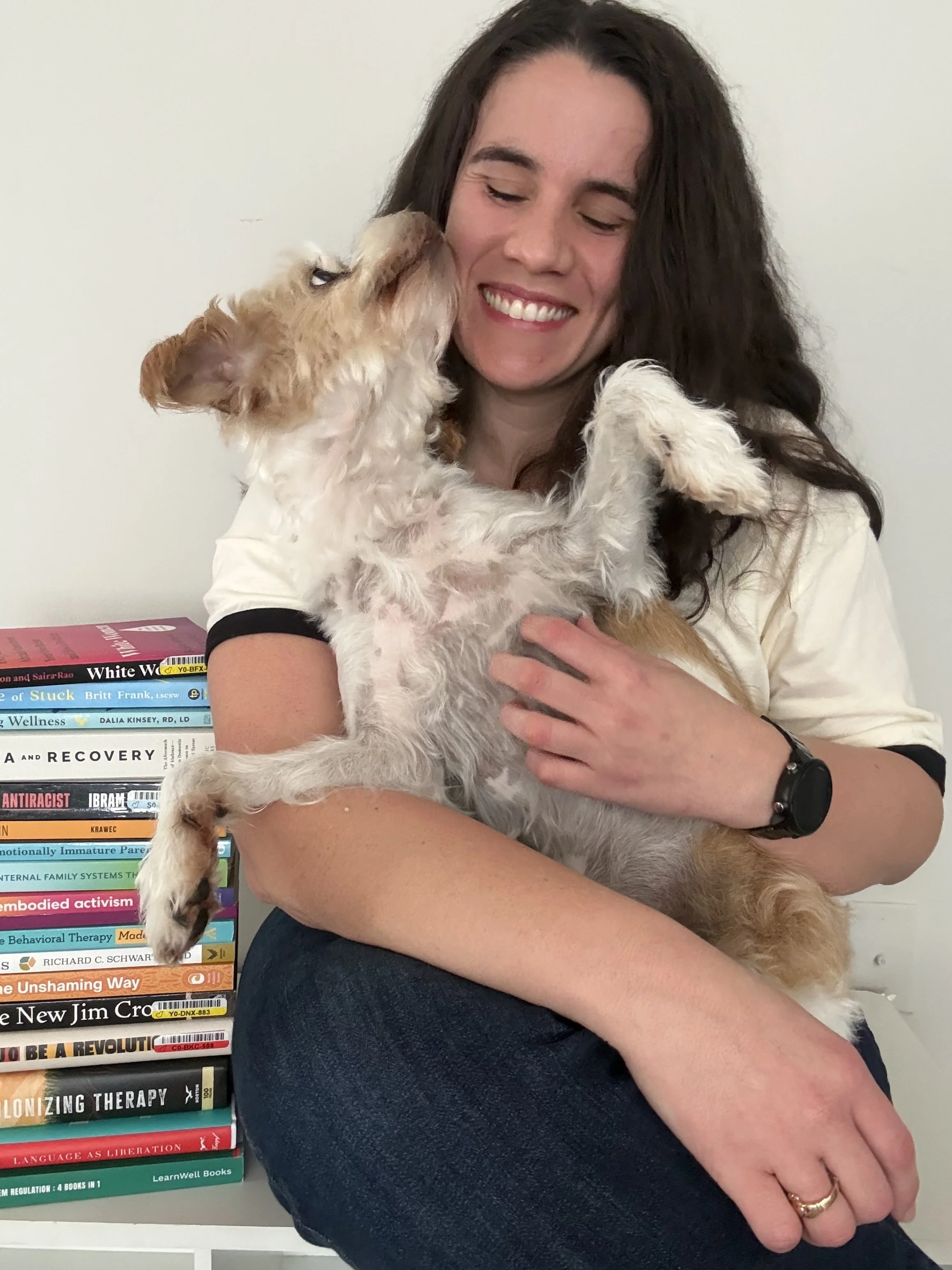 A woman with long dark hair and a watch on her wrist is sitting and holding a smiling puppy with curly fur, wearing a white shirt, in front of a stack of books.