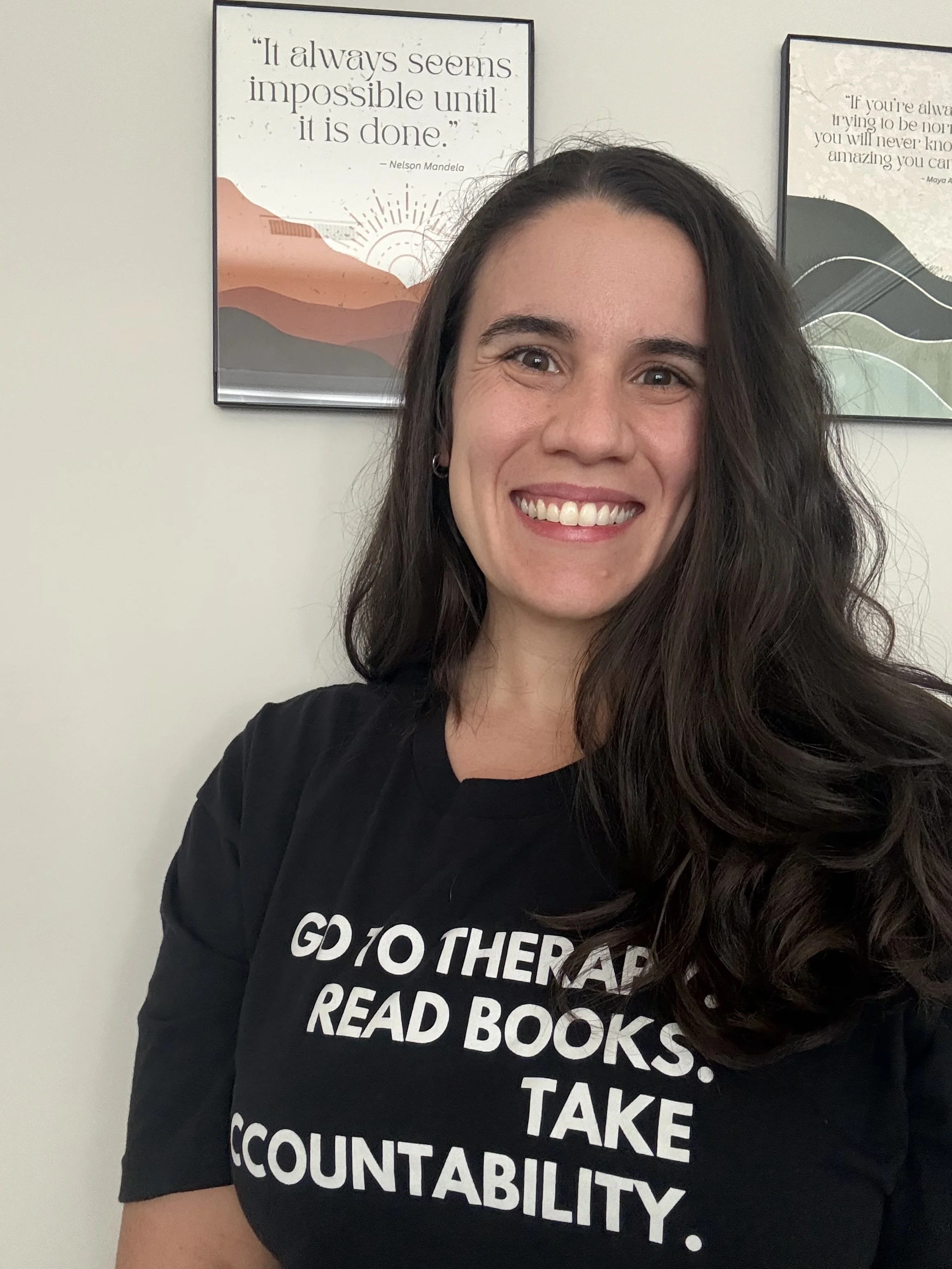 Amanda Kenney, LCSW standing in front of framed quote "It always seems impossible until it is done - Nelson Mandela," Amanda is wearing a black shirt with the words "Go to therapy, read books, take accountability"