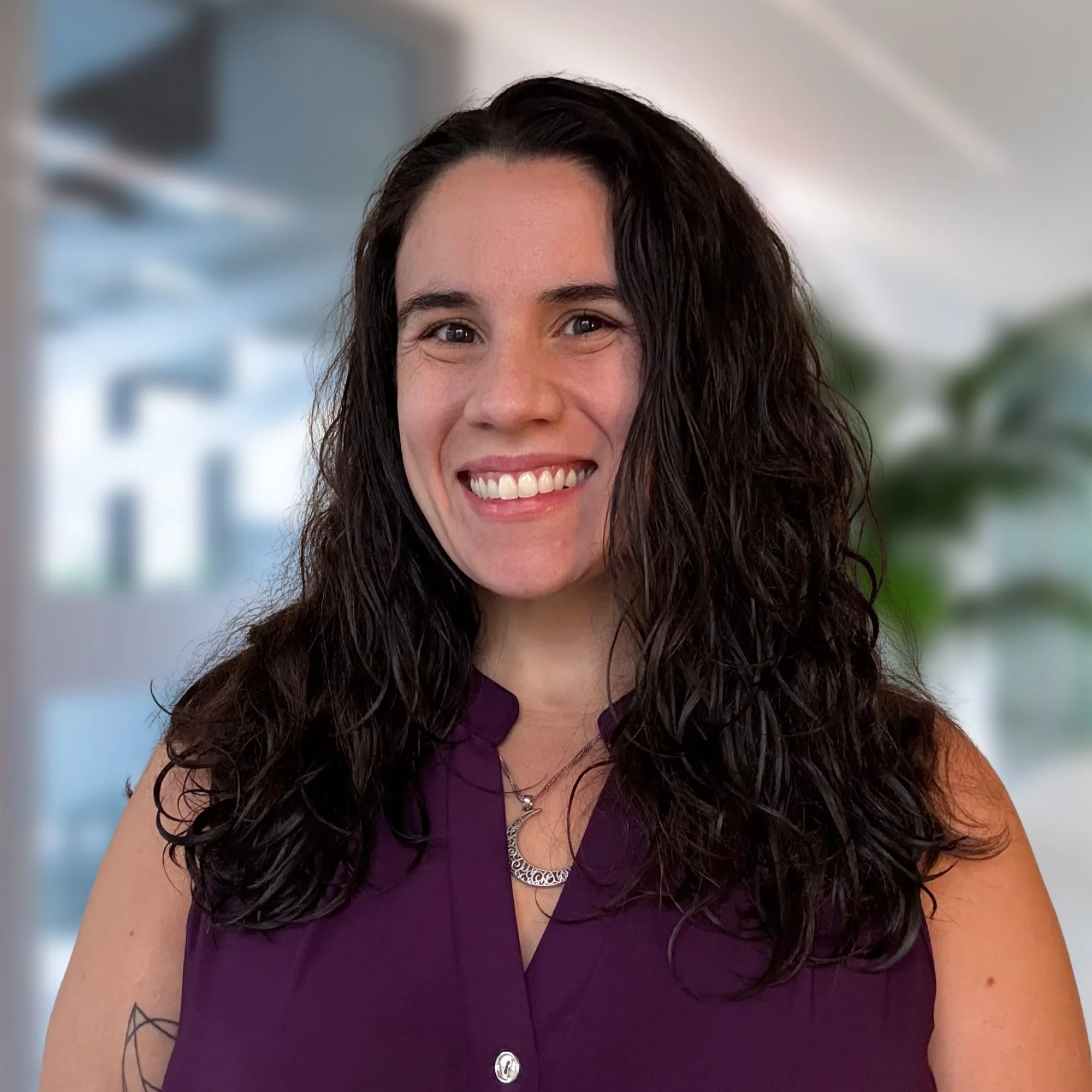 A woman with dark curly hair smiling at the camera, wearing a purple sleeveless blouse and a silver necklace, with a blurred office background.