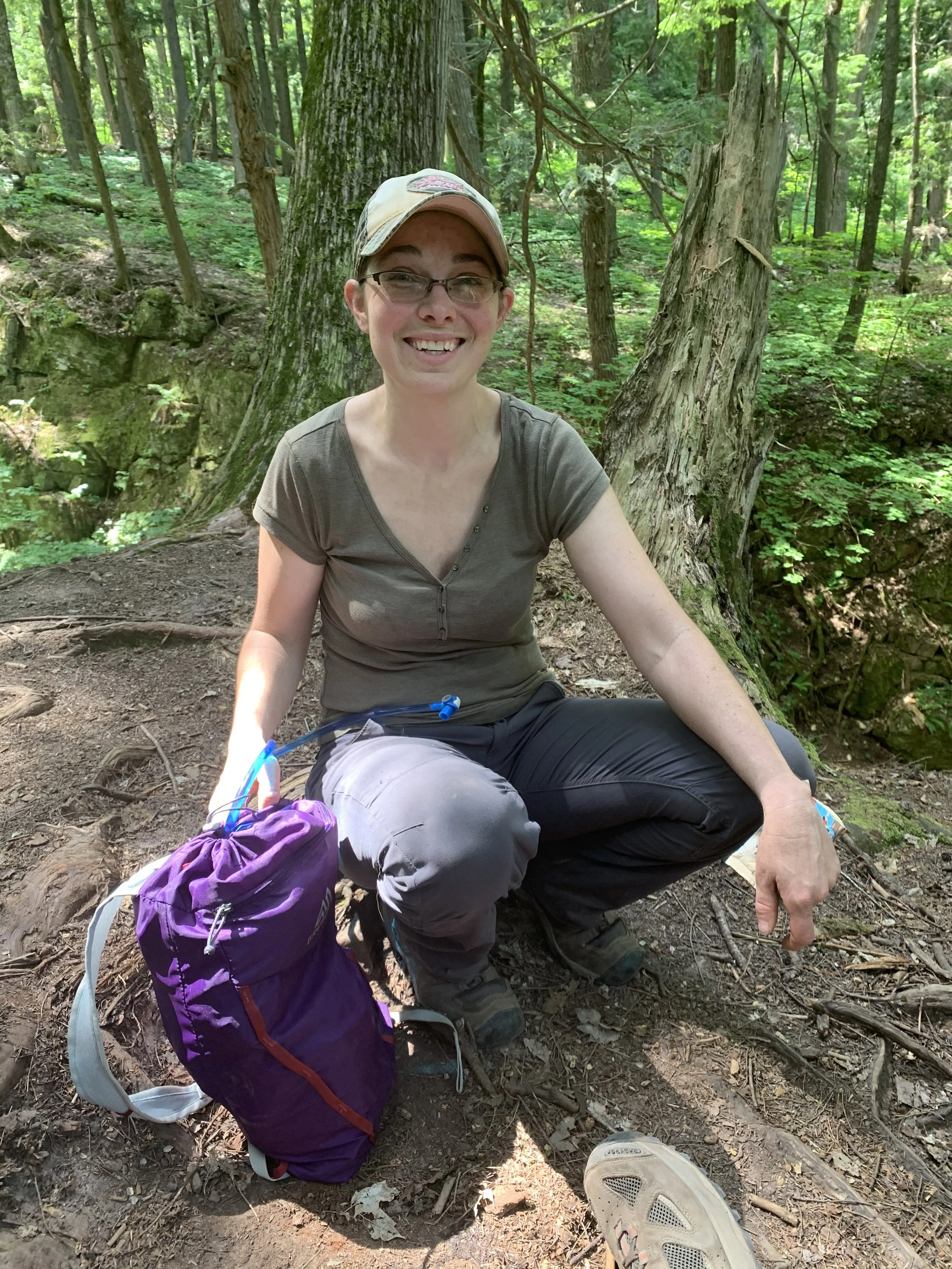 A smiling female hiker in outdoor gear crouches on a forest trail, surrounded by lush greenery, taking a moment to enjoy the adventure.