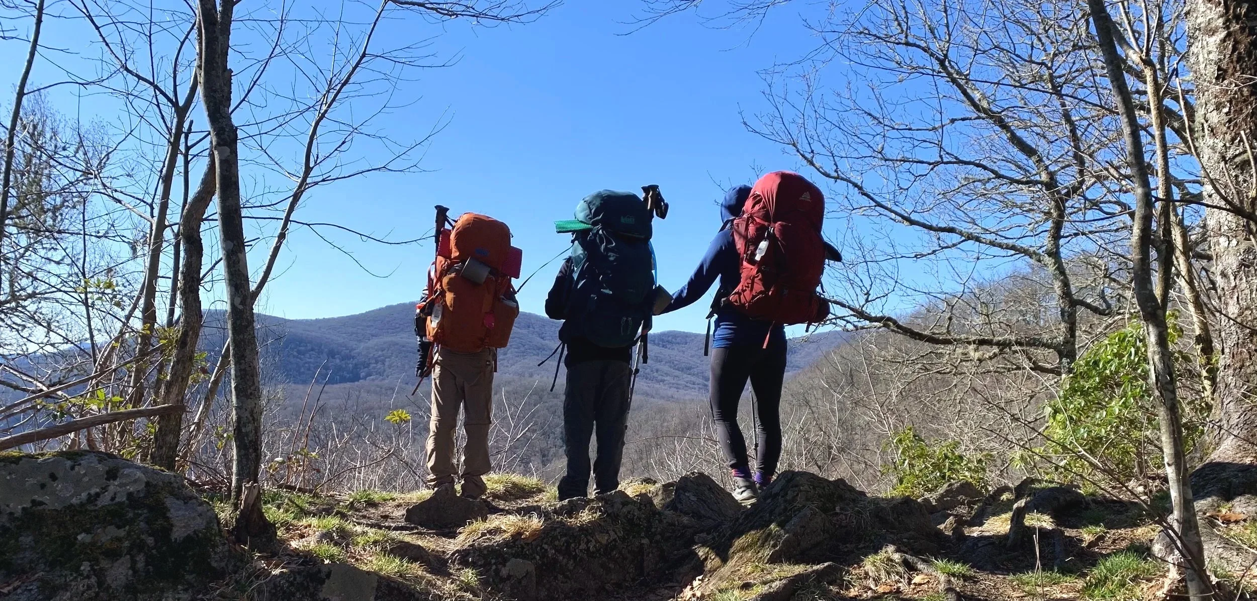 A mom and her two kids with full backpacks pause on the Appalachian Trail, taking in the mountains while trees surround them. A perfect moment of adventure.
