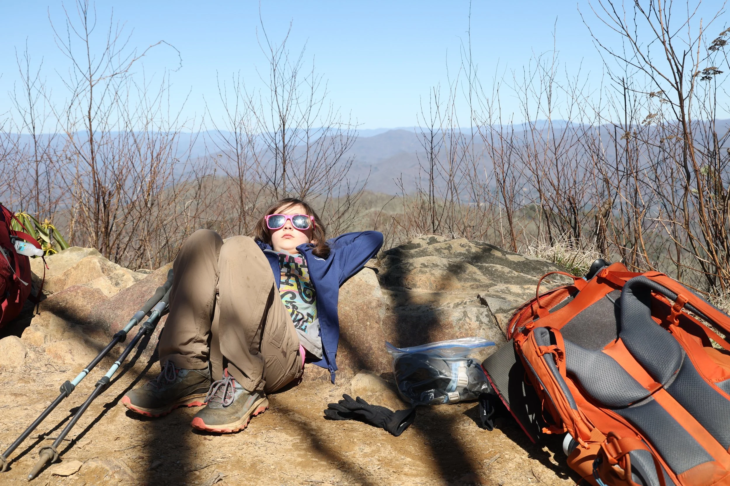 A young hiker in a blue jacket and sunglasses sprawls on a boulder, trekking poles and backpack at her side, soaking up the mountain view and enjoying a well-earned moment of trail-side relaxation.