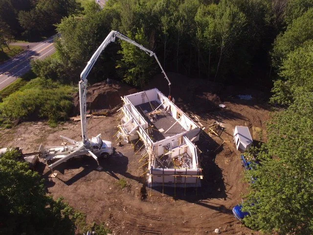 A drone view of a construction site with a house under construction, surrounded by trees, with a concrete pump truck pouring concrete into the foundation.