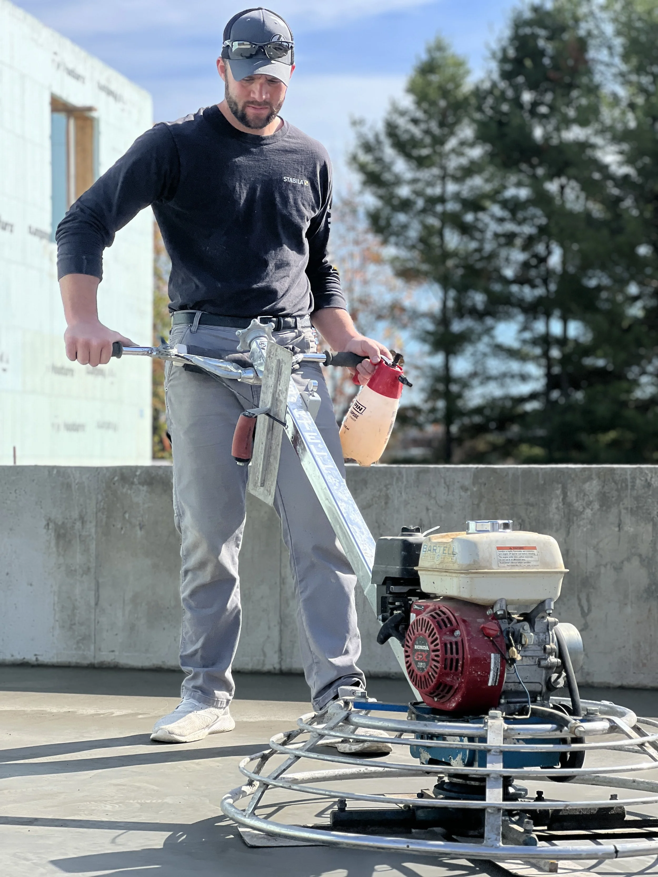 A construction worker operating a concrete power trowel on a building site, wearing sunglasses, a black long-sleeve shirt, and grey pants.