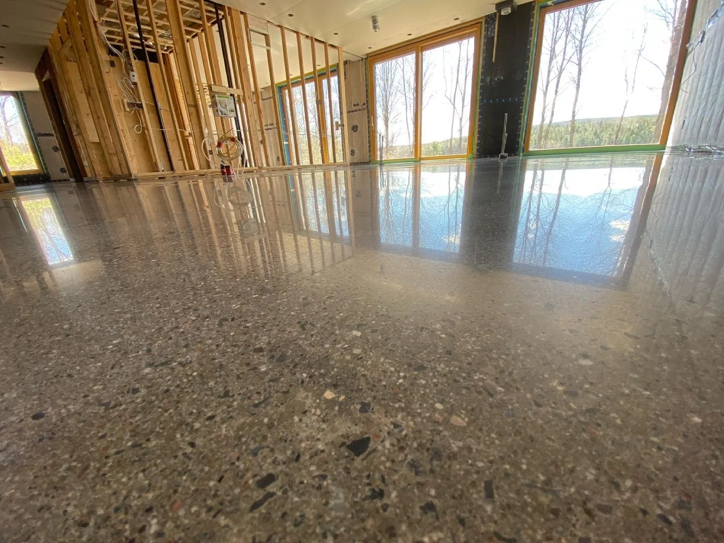 Interior of a house under construction with shiny, polished concrete floors, exposed wooden framing, and large windows showing trees outside.