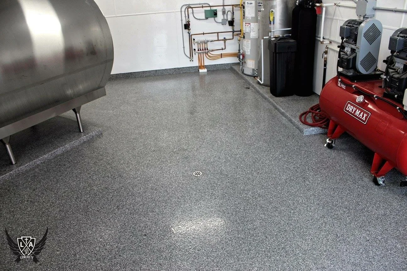 A clean, gray speckled concrete floor in an industrial or utility room, with various equipment including a stainless steel tank, a red air compressor labeled 'Dry Max,' and other machinery and pipes along the wall.