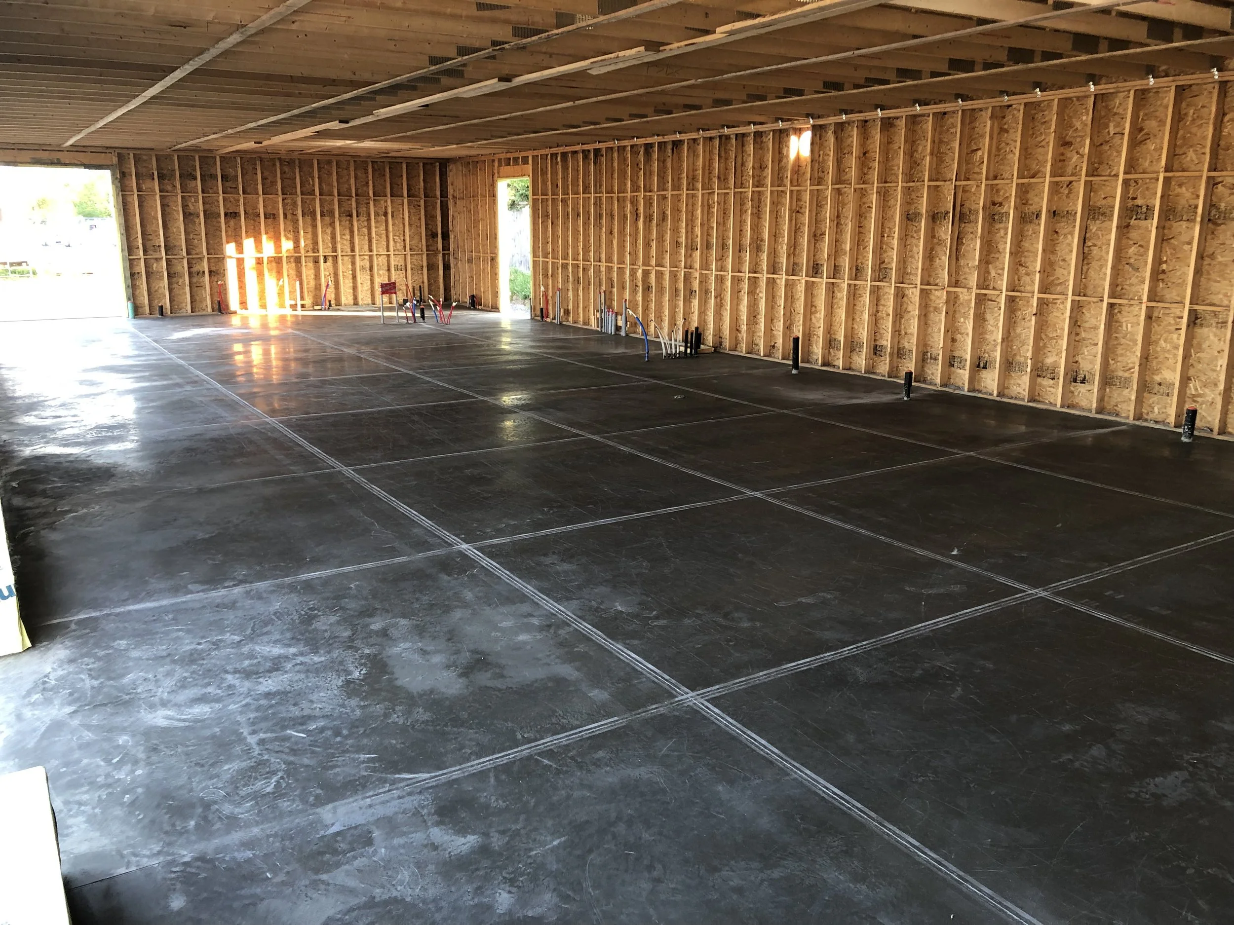 Interior of an unfinished garage with a concrete floor marked with white chalk lines for parking spaces, exposed wooden framing, and two small open doorways on the back wall.