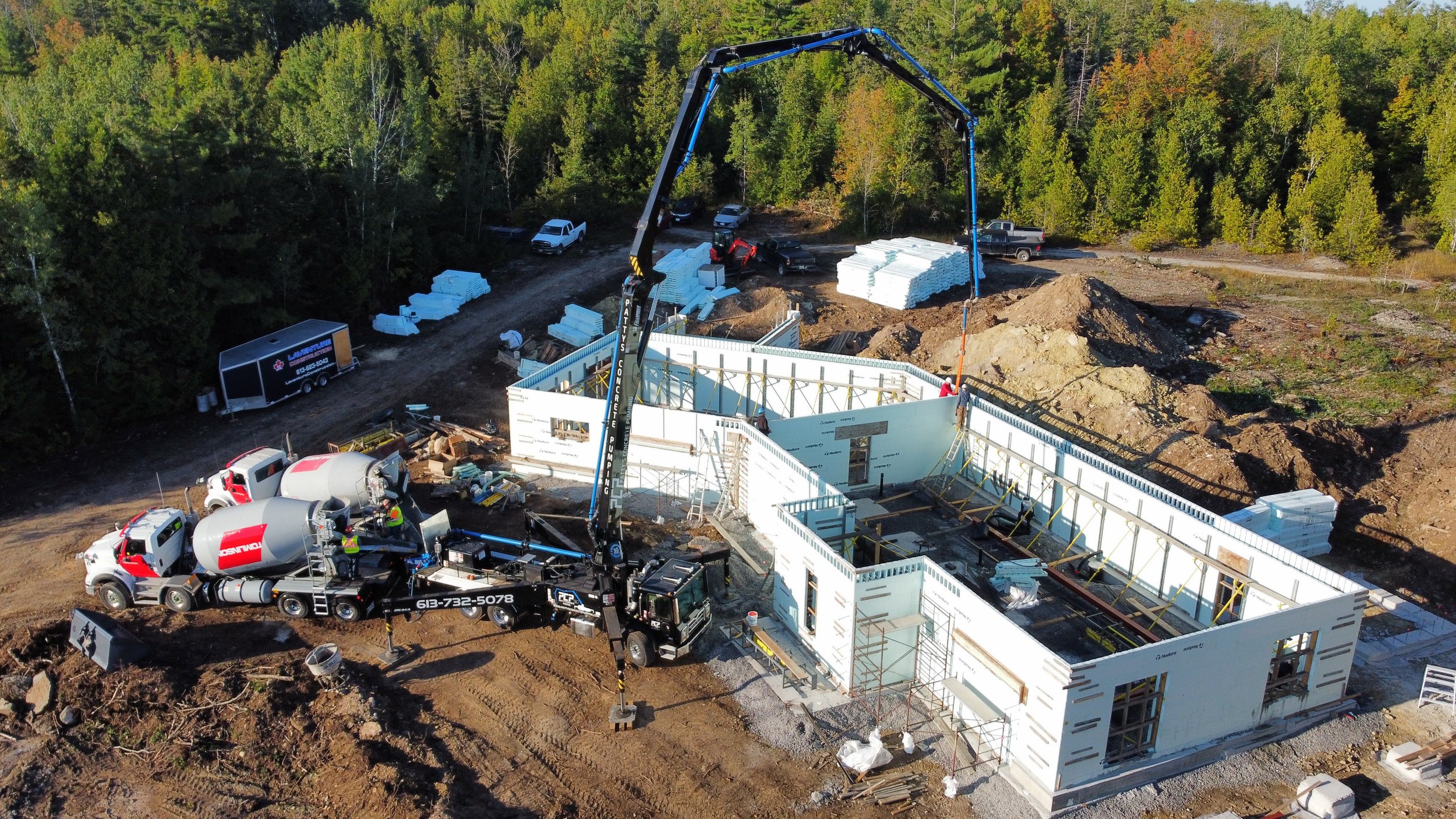 Construction site with workers and concrete mixer trucks pouring concrete into building foundation, surrounded by trees and construction materials.