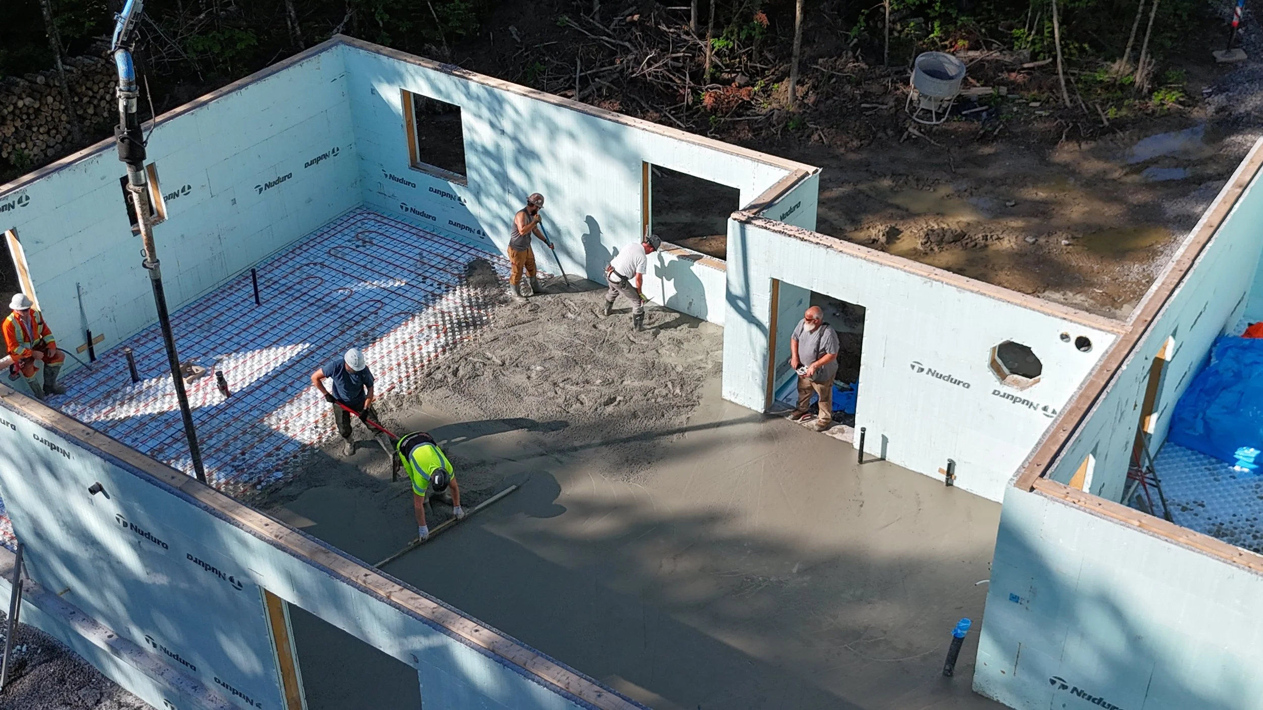 Construction workers pouring and smoothing concrete inside a building foundation, with insulated wall panels installed around the perimeter.