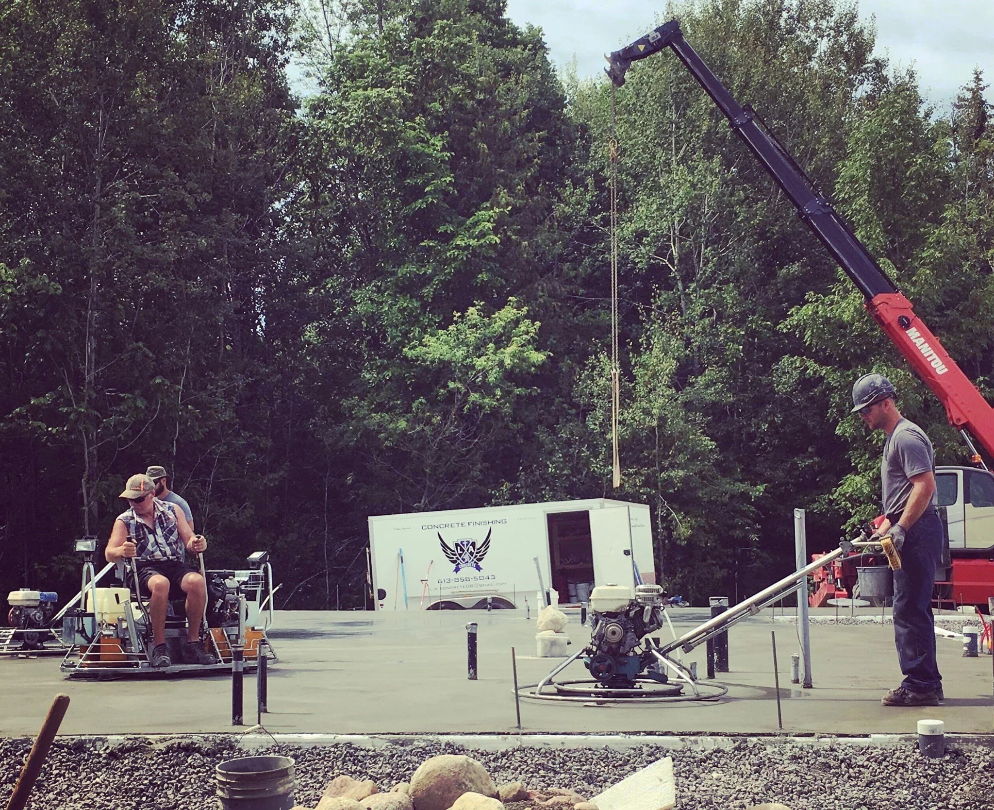 Workers pouring concrete with construction equipment, truck, and crane in a wooded area.