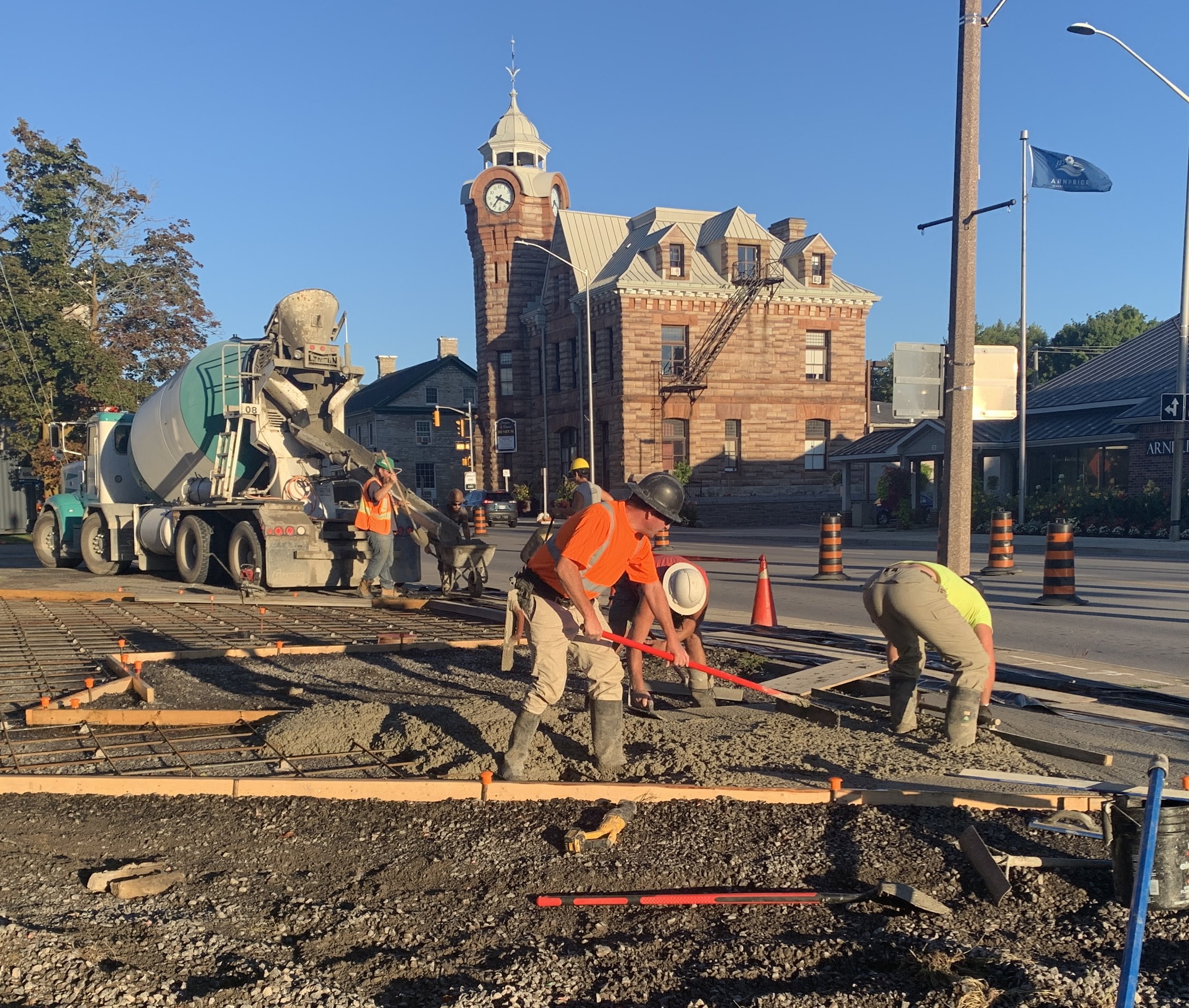 Construction workers laying concrete on a city street at sunset, with a historic brick building and a clock tower in the background.