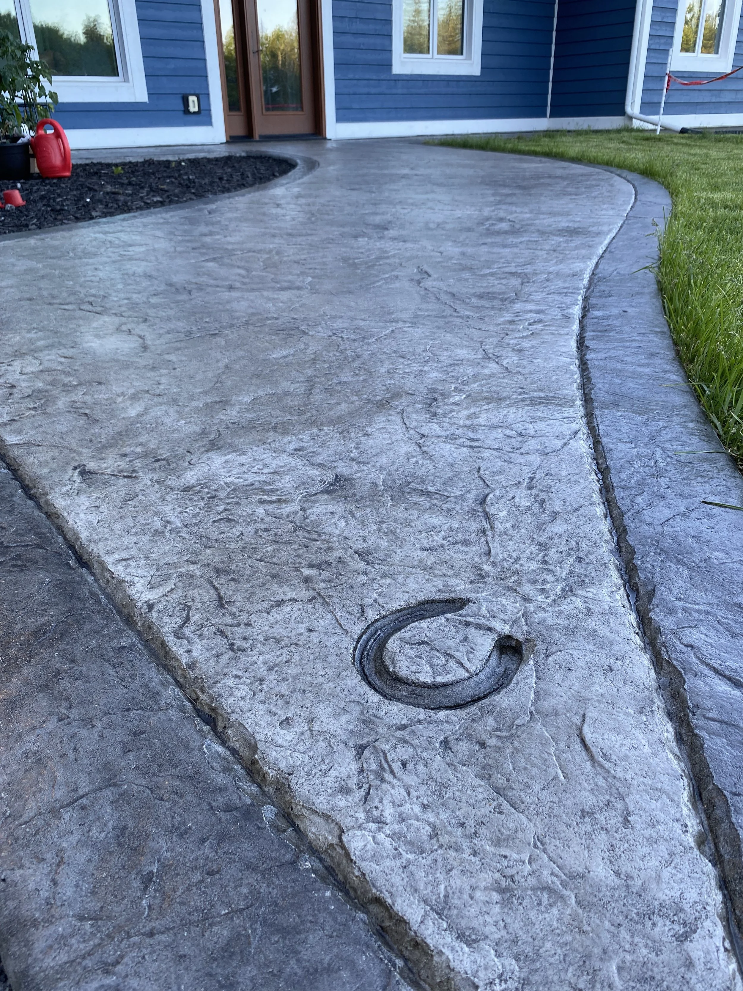 Close-up of a newly poured concrete sidewalk with expansion joints and a metal utility cover, in front of a blue house with white trim.