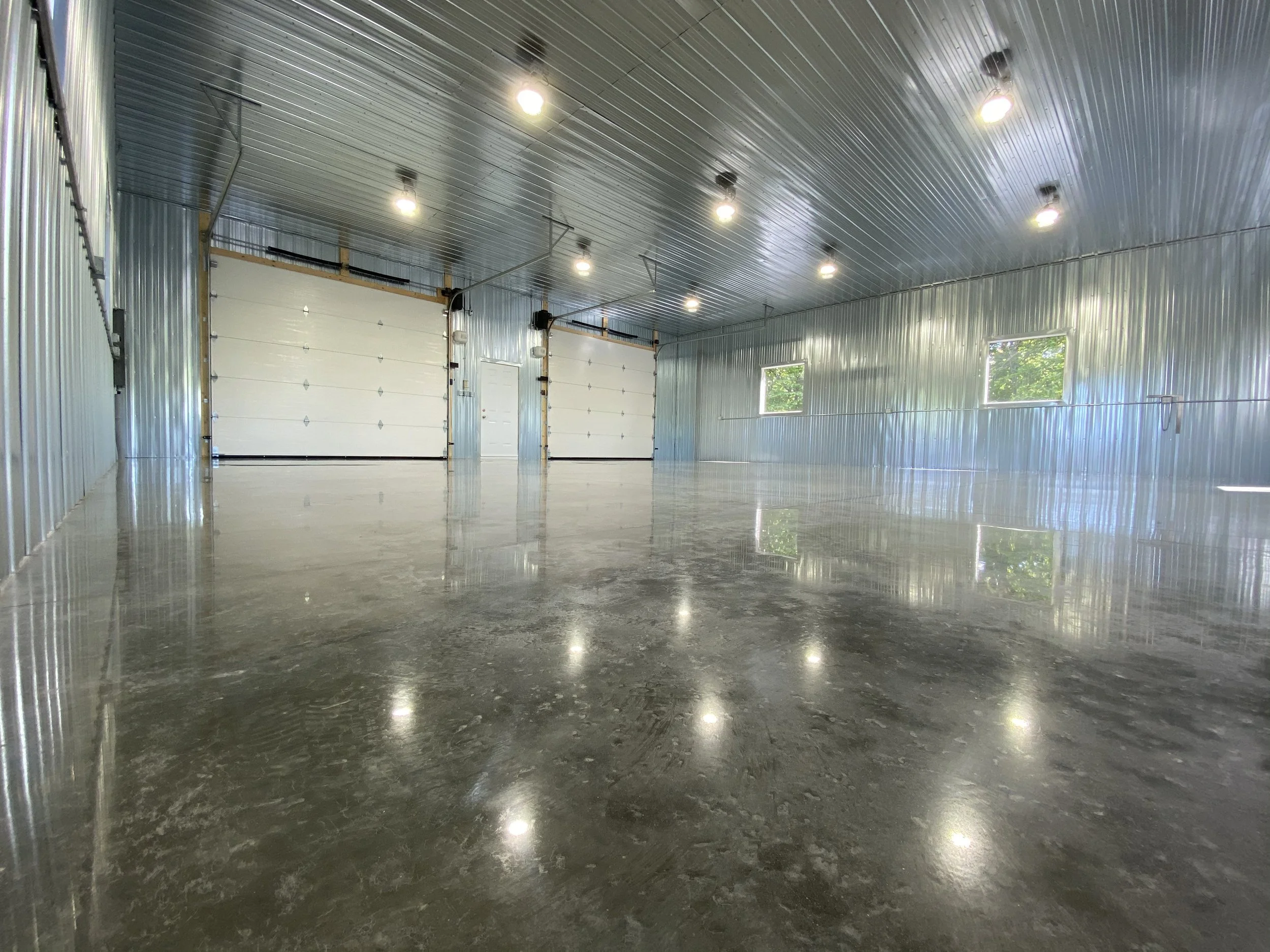 Empty, metallic interior garage with polished concrete floor, metal walls and ceiling, fluorescent lighting, two large garage doors, a side door, and small windows near the ceiling reflecting the interior lights.