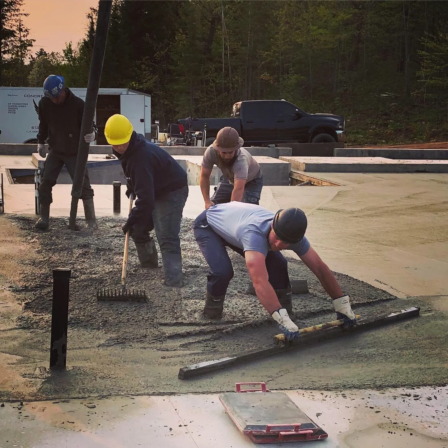 Four workers in helmets and gloves pouring and spreading concrete on a construction site, with trucks and vehicles in the background and trees beyond.