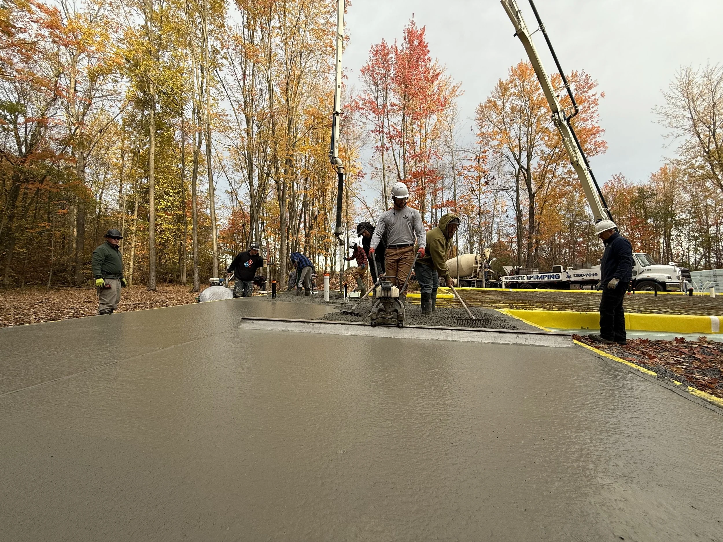 Construction workers are pouring and smoothing concrete on a new sidewalk in a wooded area with fall foliage.