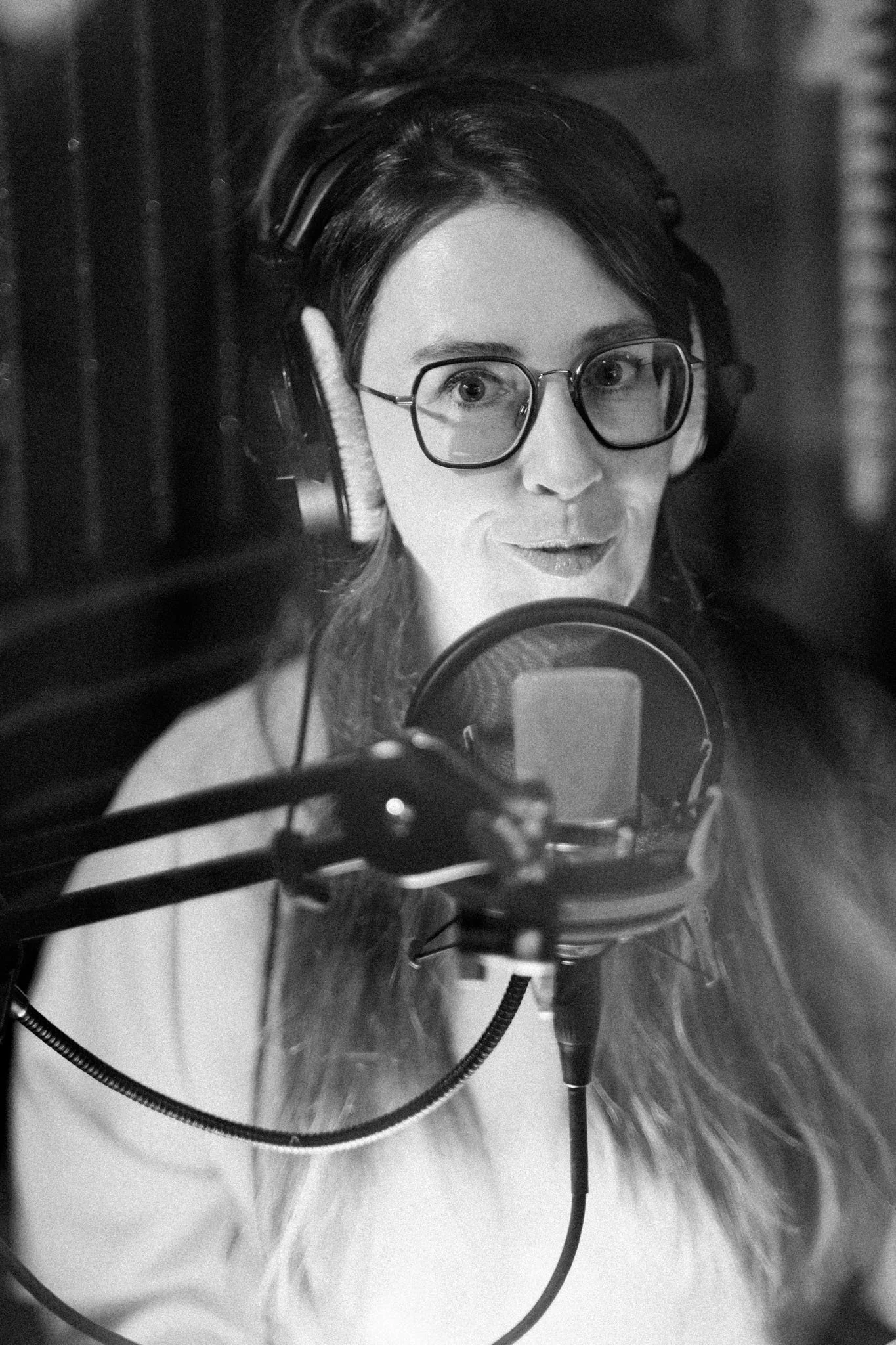 A woman with glasses and headphones speaking into a microphone in a recording studio.