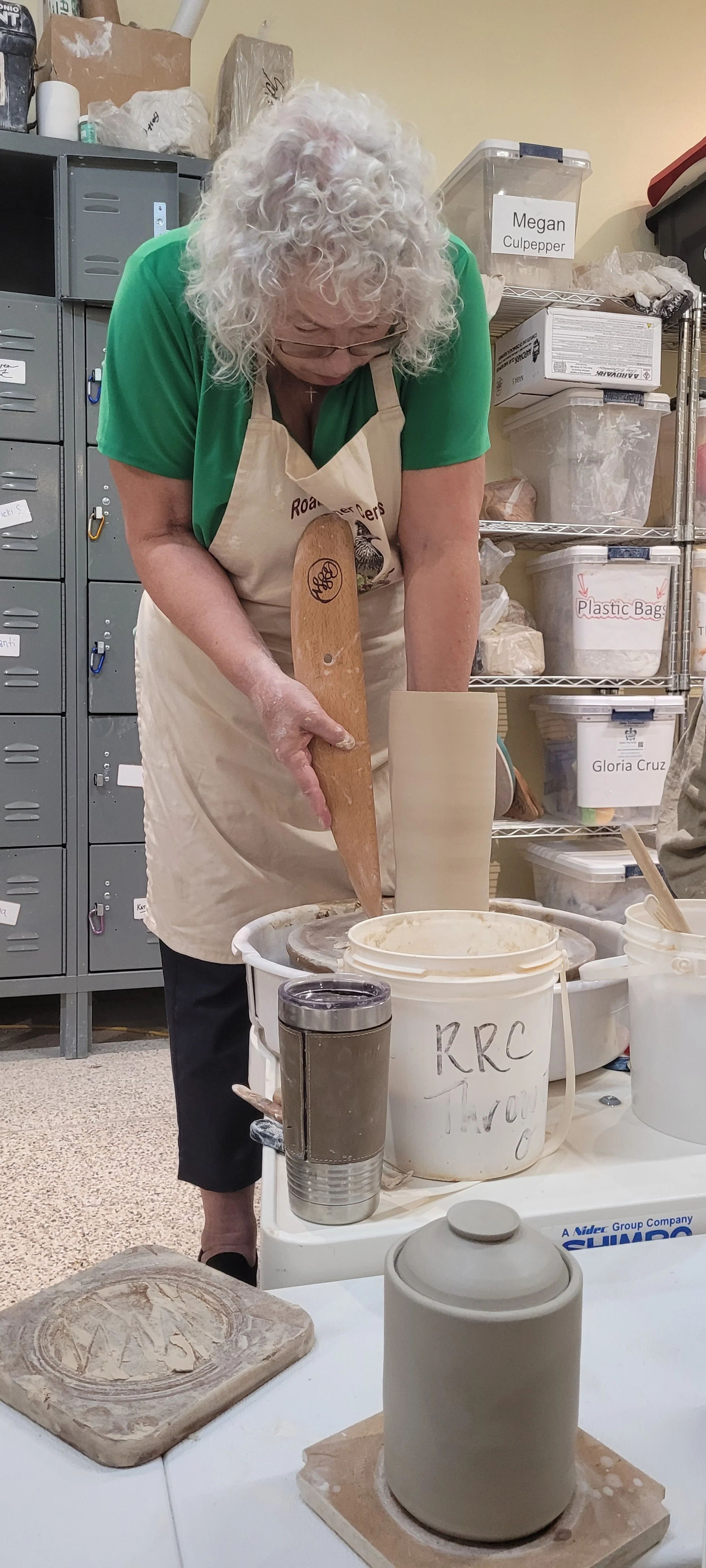 A woman with curly gray hair and glasses working with clay in a pottery studio. She is wearing a beige apron and green shirt, and is using a pottery wheel to shape a tall vase. Various tools and containers are visible around her.