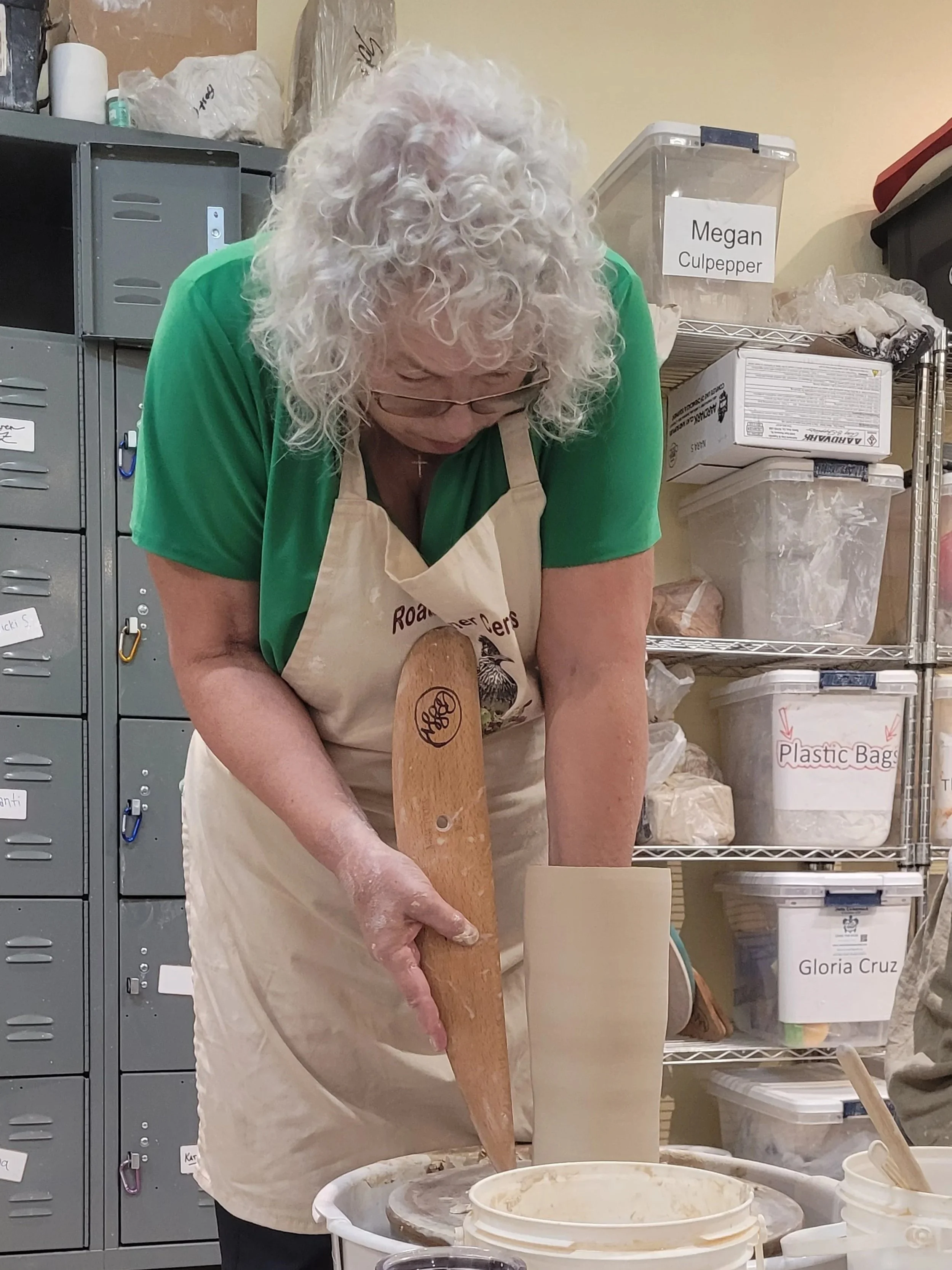 A woman with curly white hair and glasses, wearing a green shirt and a beige apron, is working in a ceramics or pottery studio. She is holding a wooden paddle and appears to be shaping or working on a pottery piece on a wheel, with various supplies a