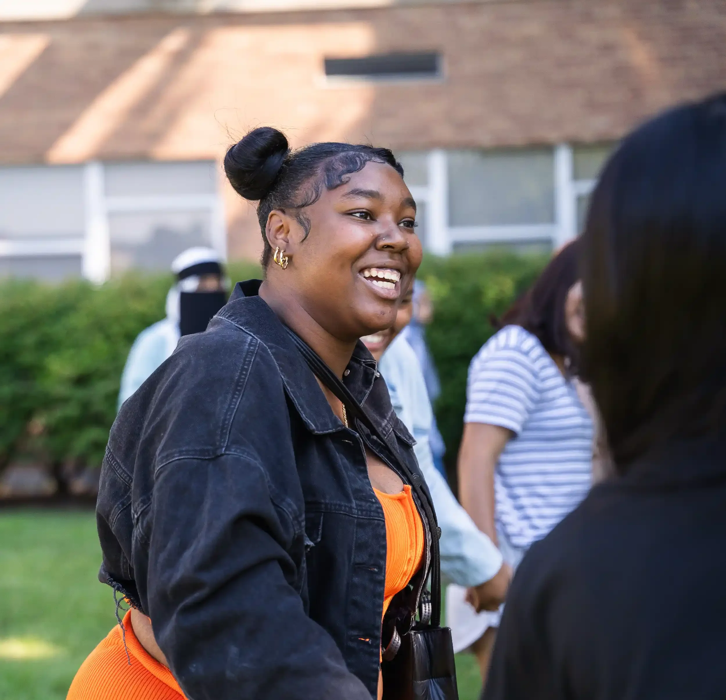 A young woman smiling and talking to someone outdoors, wearing a black jacket and orange dress, with people and a building in the background.