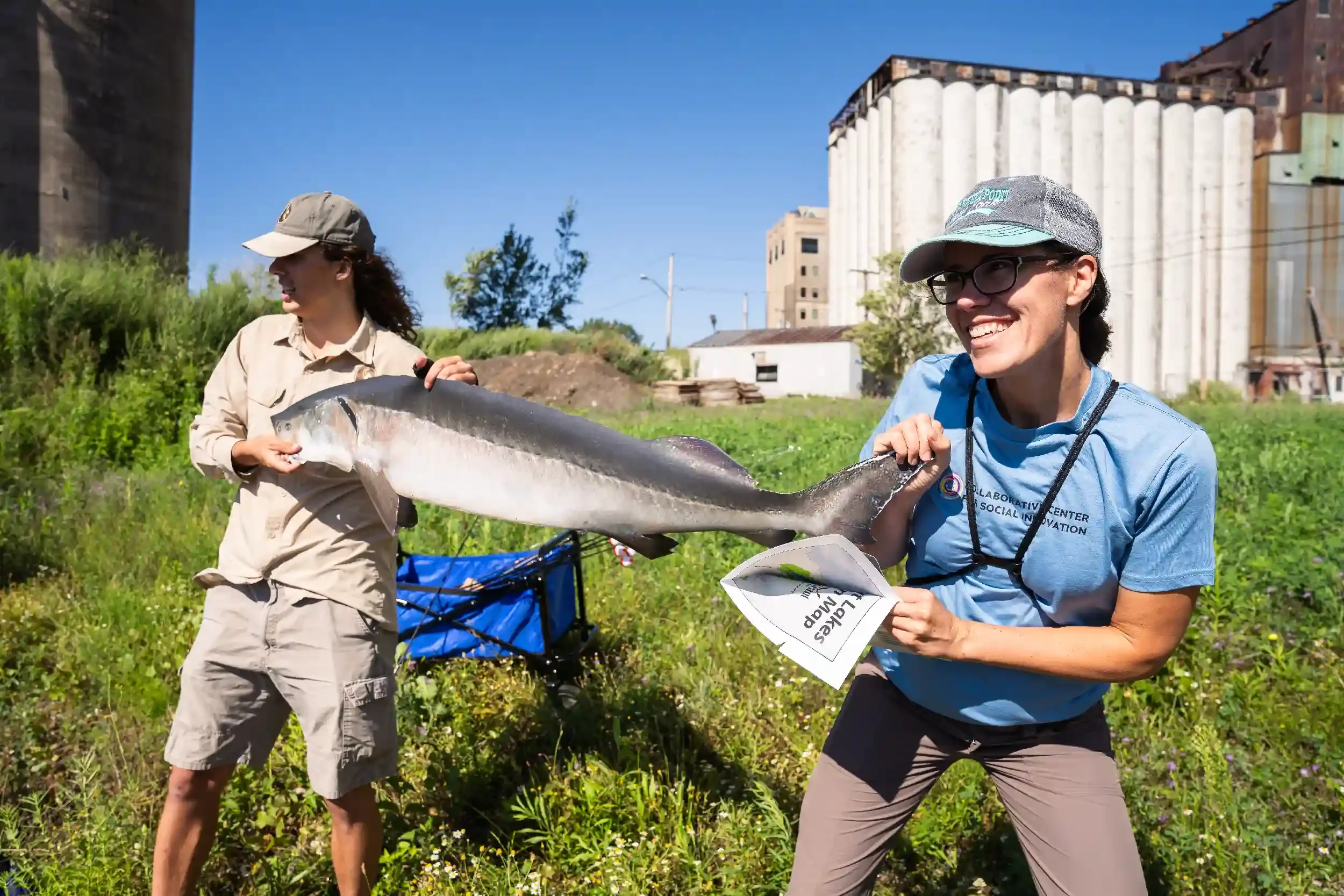 Two women outdoors holding a large fish, one in the foreground smiling and wearing a cap and glasses, the other in the background looking at the fish, with green grass and industrial buildings in the background.