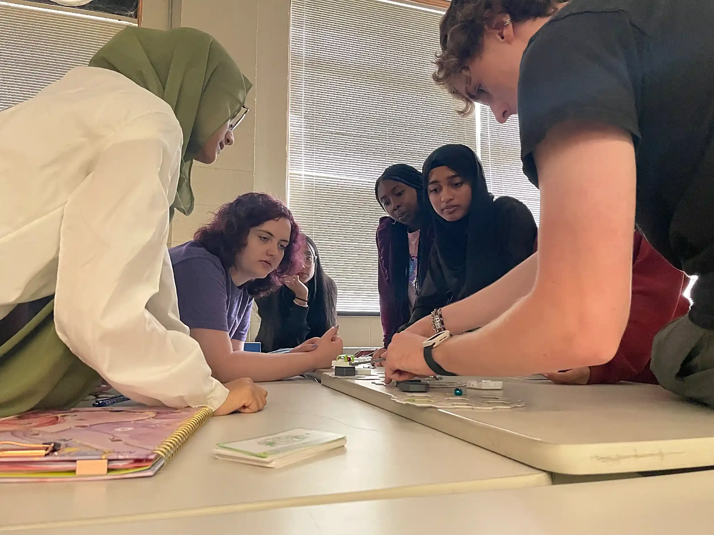 A group of diverse people gathered around a table, watching and engaging with a game or activity on the table, in a room with blinds covering a window.