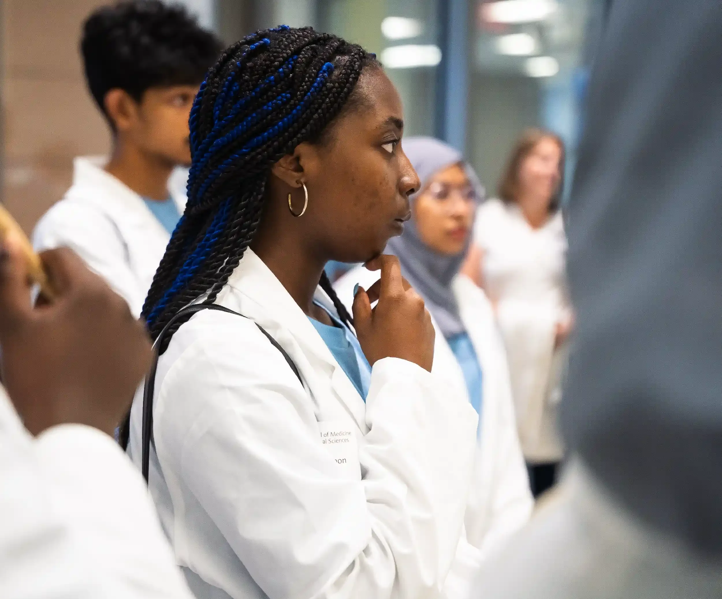 A diverse group of medical students wearing white lab coats, standing in a line, with one woman in the foreground deep in thought, with her hand on her chin, during a professional training session.