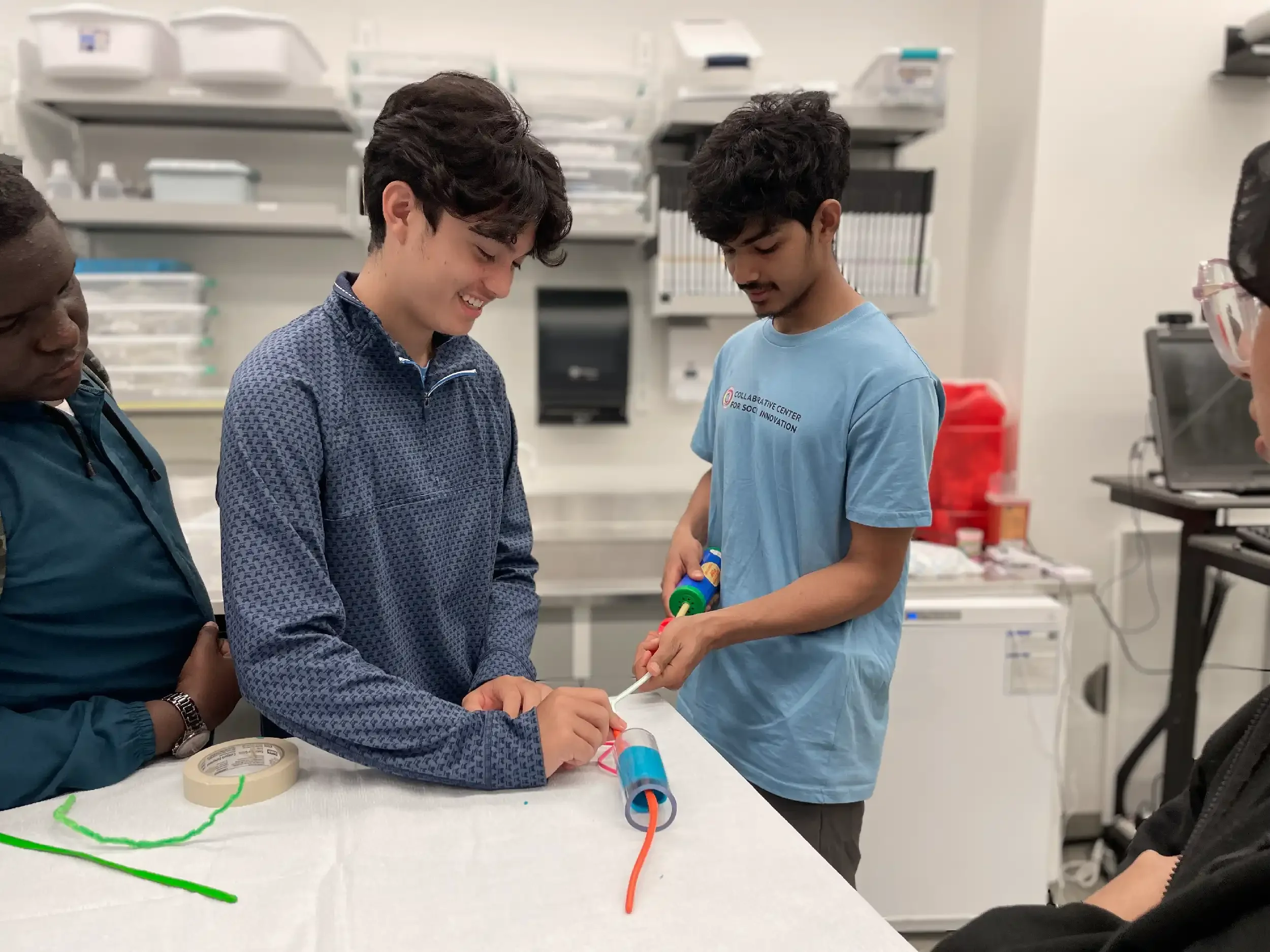 A group of young people in a laboratory creating a simple circuit with colorful wires and a battery holder, while smiling and observing their work.