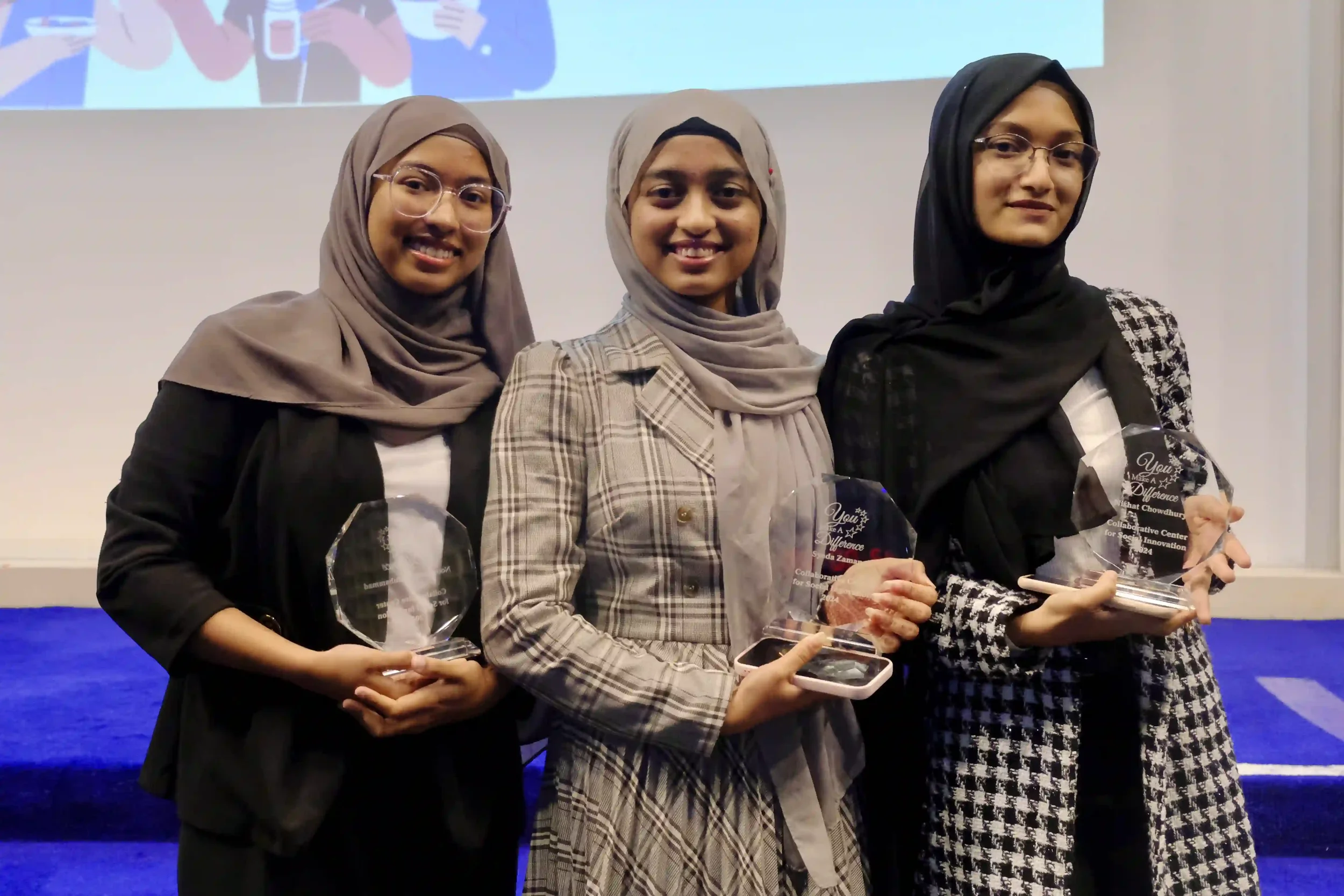 Three women wearing hijabs and professional attire holding awards at an indoor event.