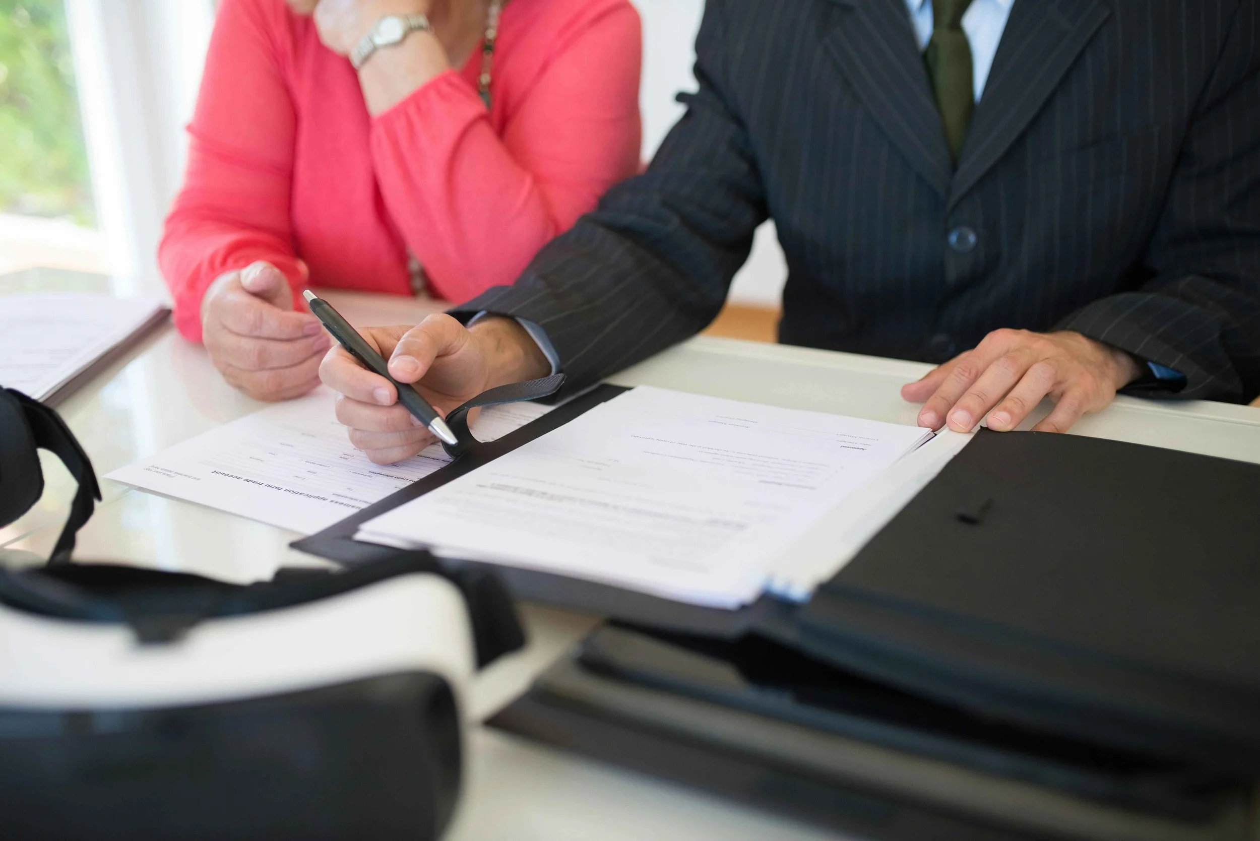 Two people sitting at a desk reviewing documents, one person in a pinstripe suit holding a pen, with a black folder and a black and white device on the desk.