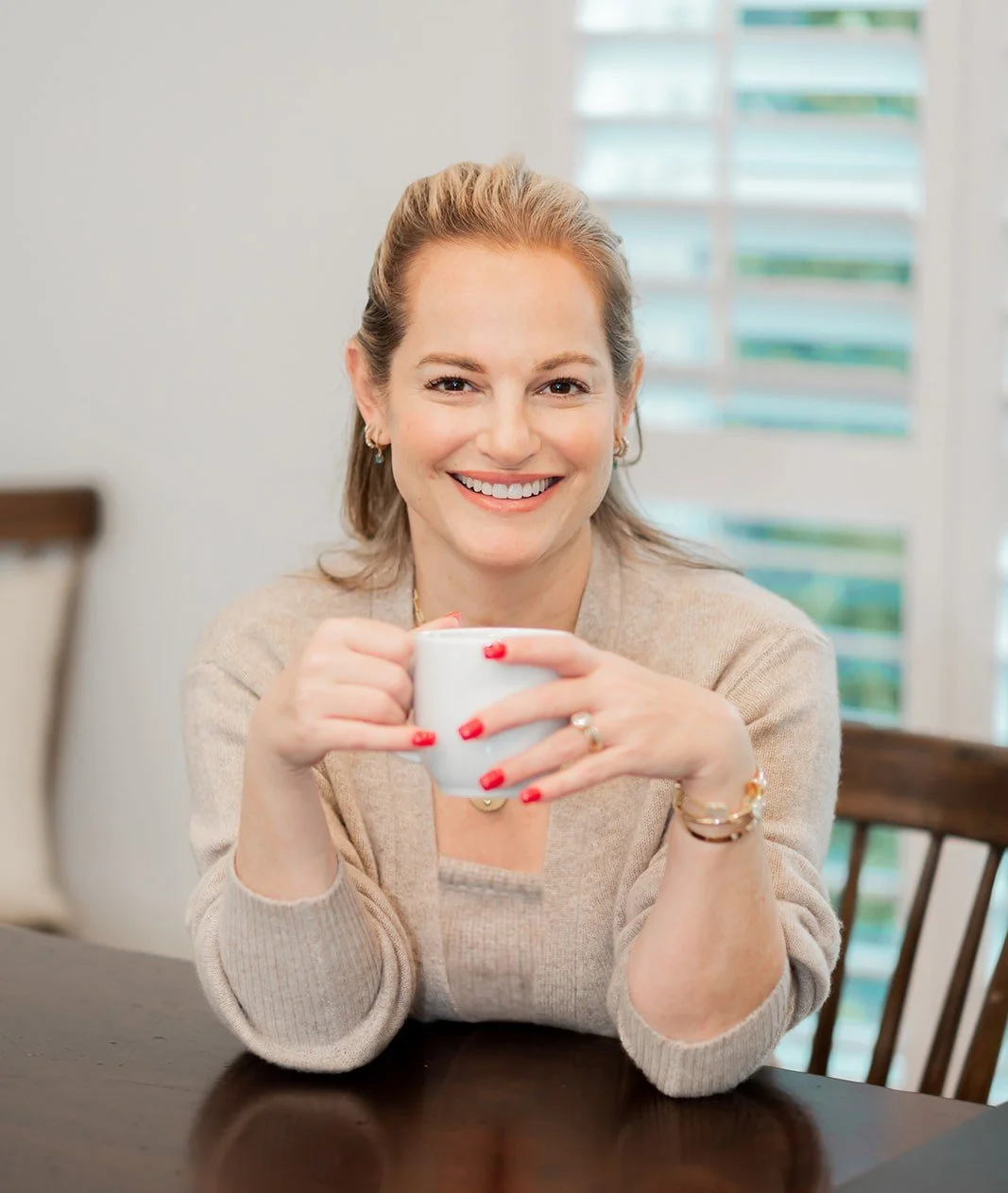 A woman with blonde hair smiling and holding a white mug, sitting at a dark wooden table in a bright room with white blinds.
