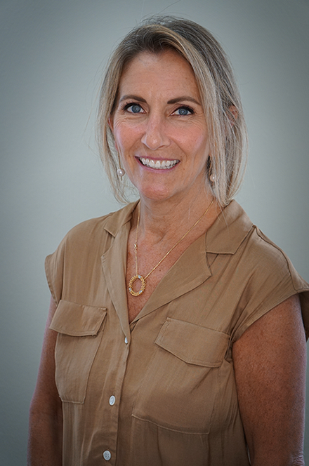 Portrait of a smiling woman with blonde hair, wearing a tan blouse and a gold necklace, standing against a plain gray background.