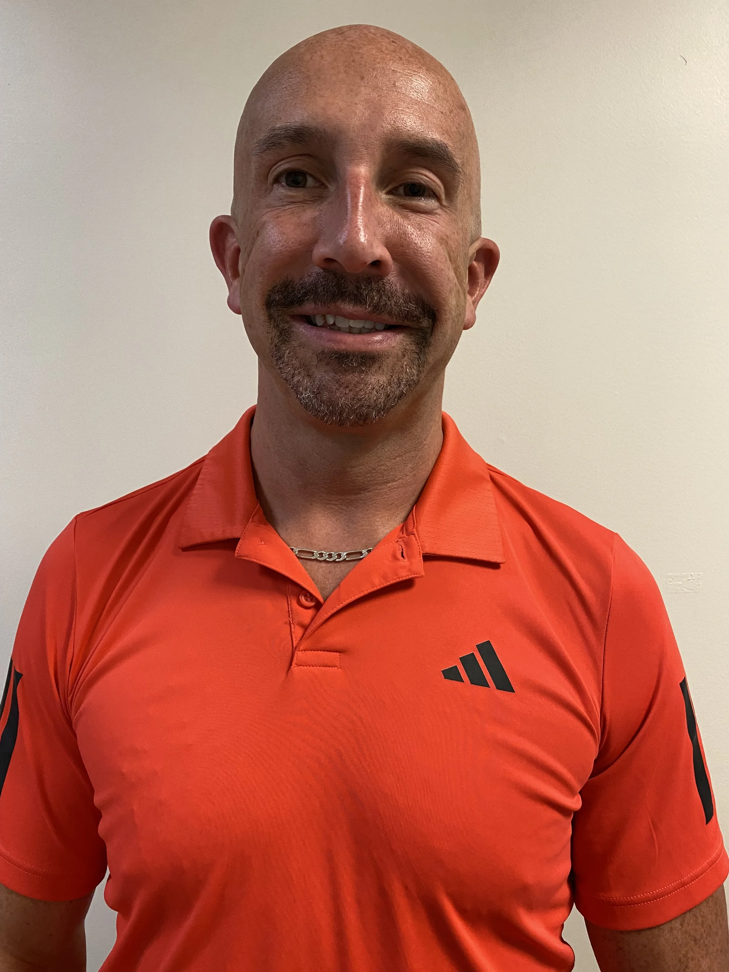 A smiling man with a bald head, goatee, and mustache wearing an orange polo shirt with black logos, standing against a plain white wall.