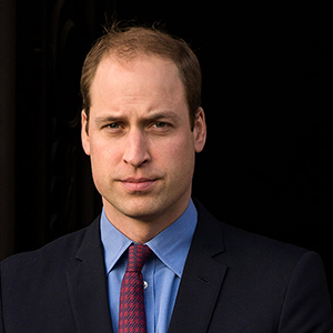 A man with short brown hair, wearing a dark suit, blue shirt, and red tie, standing outdoors against a dark background.