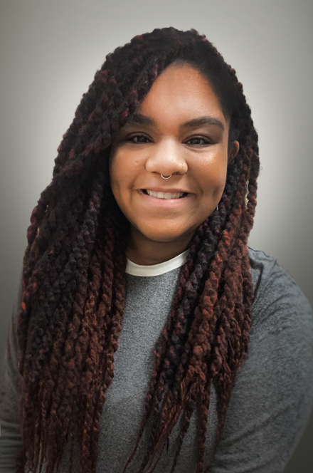 A young woman with dark skin and long, thick, reddish-brown twisted dreadlocks smiles at the camera against a plain gray background.