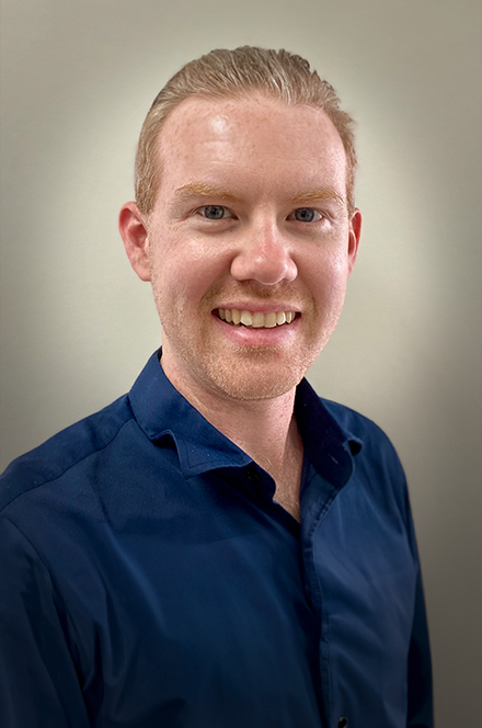 A smiling young man with light skin, short red hair, and blue eyes, wearing a navy blue dress shirt, against a plain neutral background.