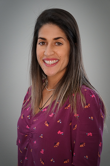 A woman with shoulder-length brown hair, smiling, wearing a purple blouse with a colorful butterfly pattern, and a silver necklace, posed against a plain gray background.