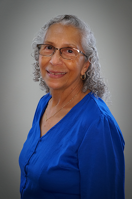 A smiling older woman with glasses and gray curly hair, wearing a blue blouse, standing against a neutral background.