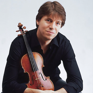 A man with brown hair holding a violin, sitting against a plain light background.